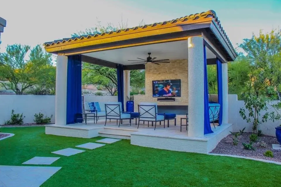 Tile-roof cabana with curtains, outdoor TV and stacked stone wall beside artificial turf in Tucson.