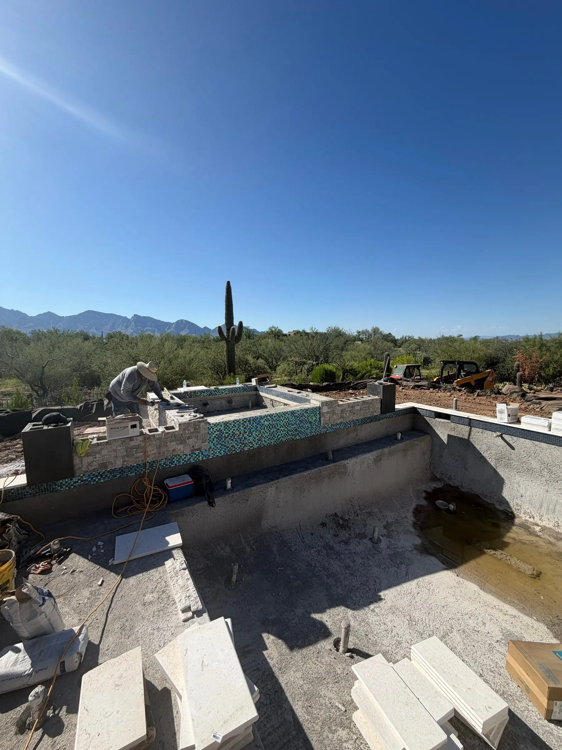 Mason setting stone veneer on a glass-tiled spa wall with coping stones staged nearby