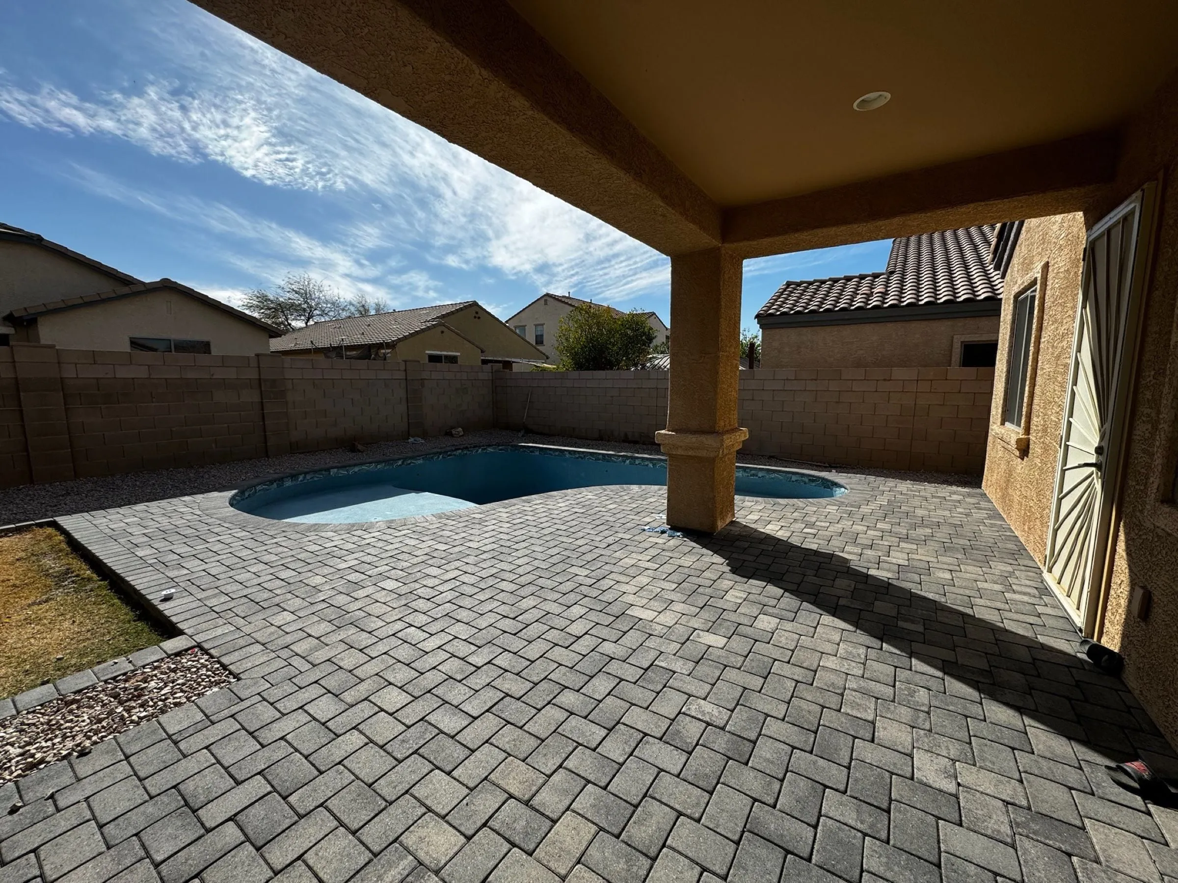 View from a covered patio across a gray herringbone paver deck to a newly filled freeform pool.