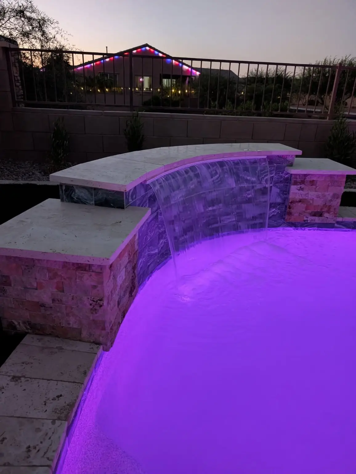 Curved raised wall with sheer descent waterfall, stacked stone and purple pool lighting at twilight.
