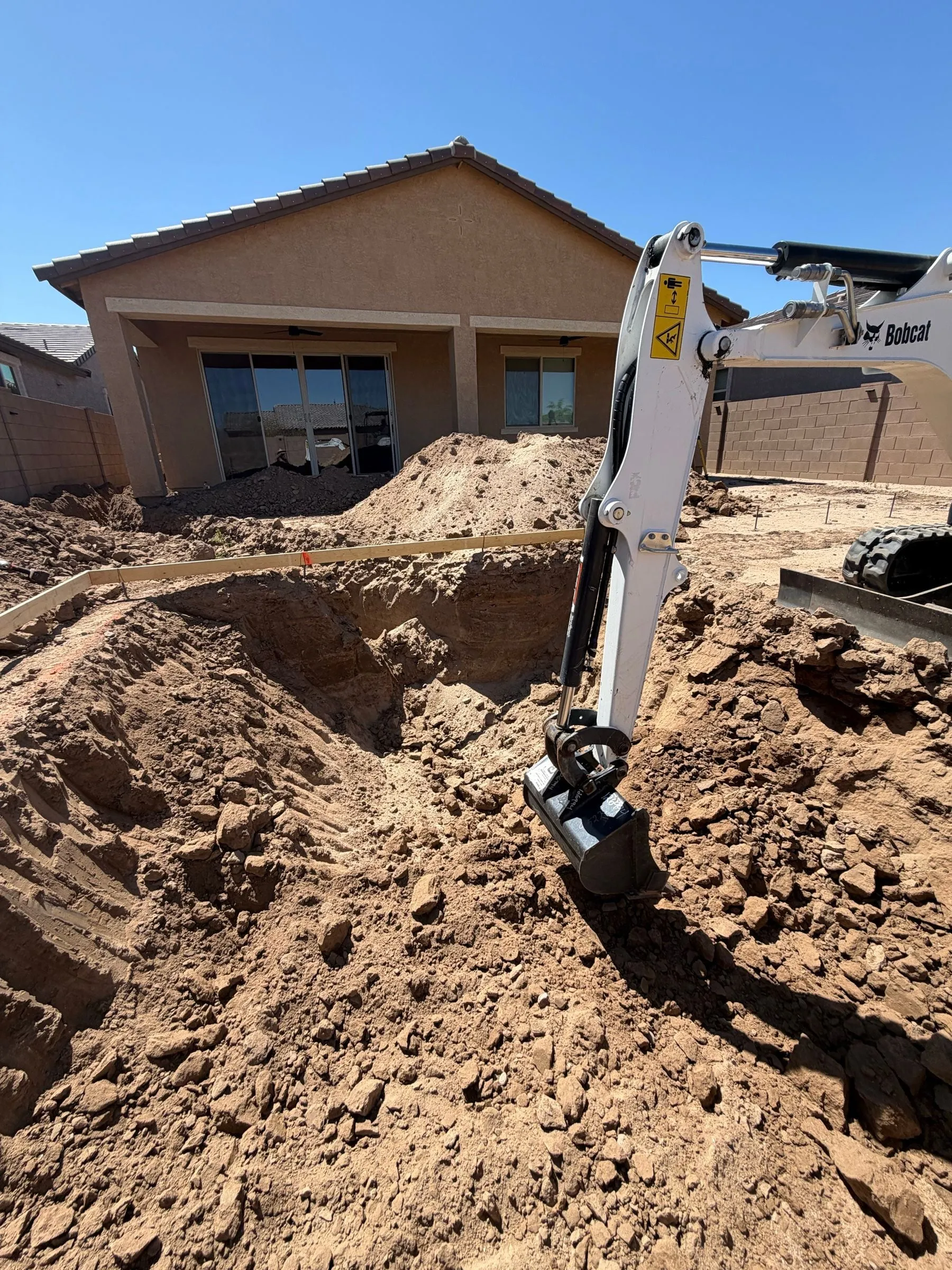 Excavator bucket digging a new pool in a dirt backyard behind a home.