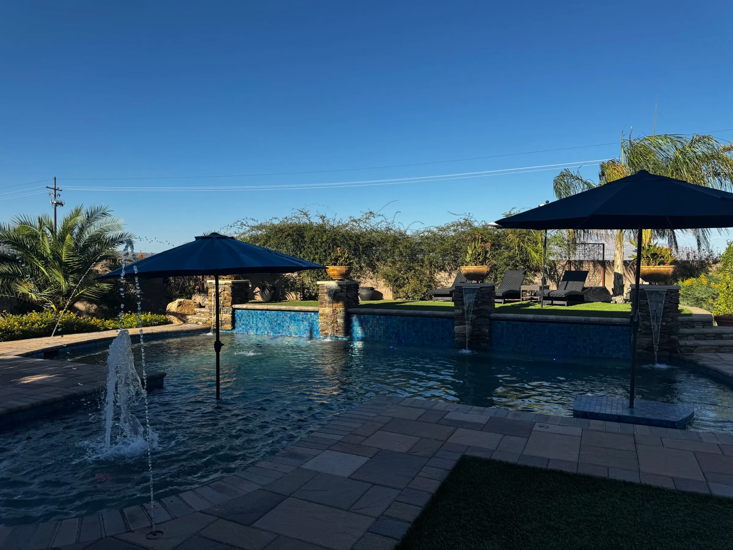 Finished freeform pool with umbrellas, bubblers, a glass tile wall and stone pillar scuppers.