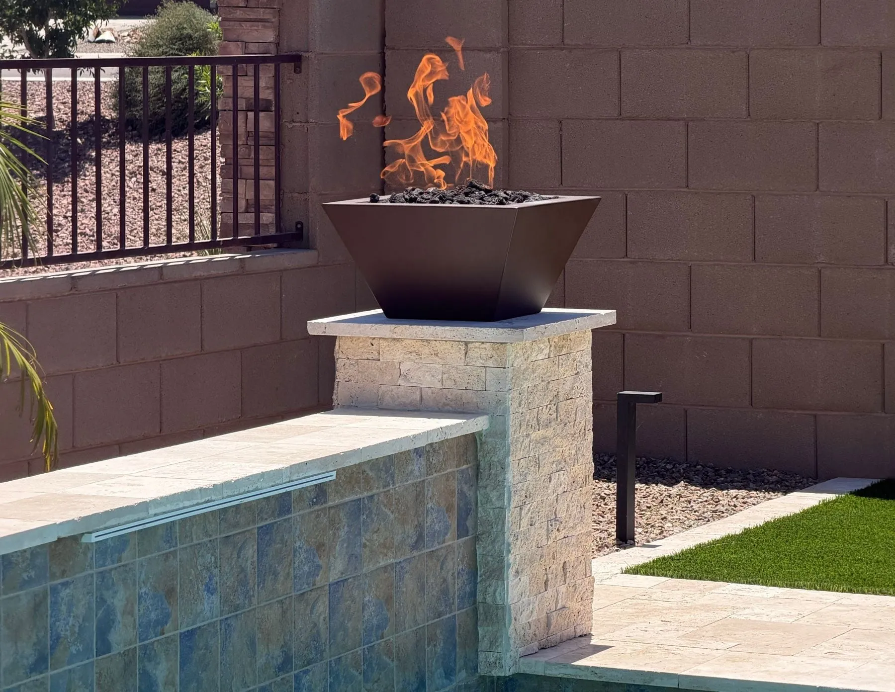 Lit fire bowl atop a stacked travertine pillar at the end of a tiled raised pool wall.