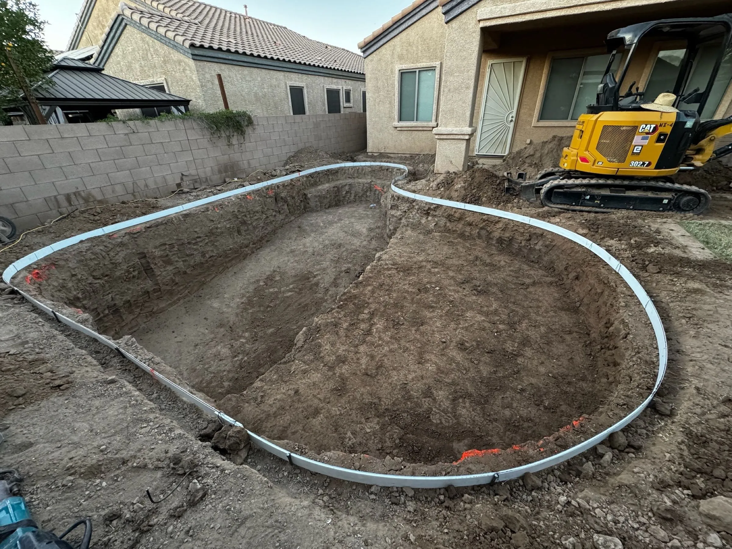 Freeform pool excavation outlined with white forming boards in Marana, with a compact excavator nearby.