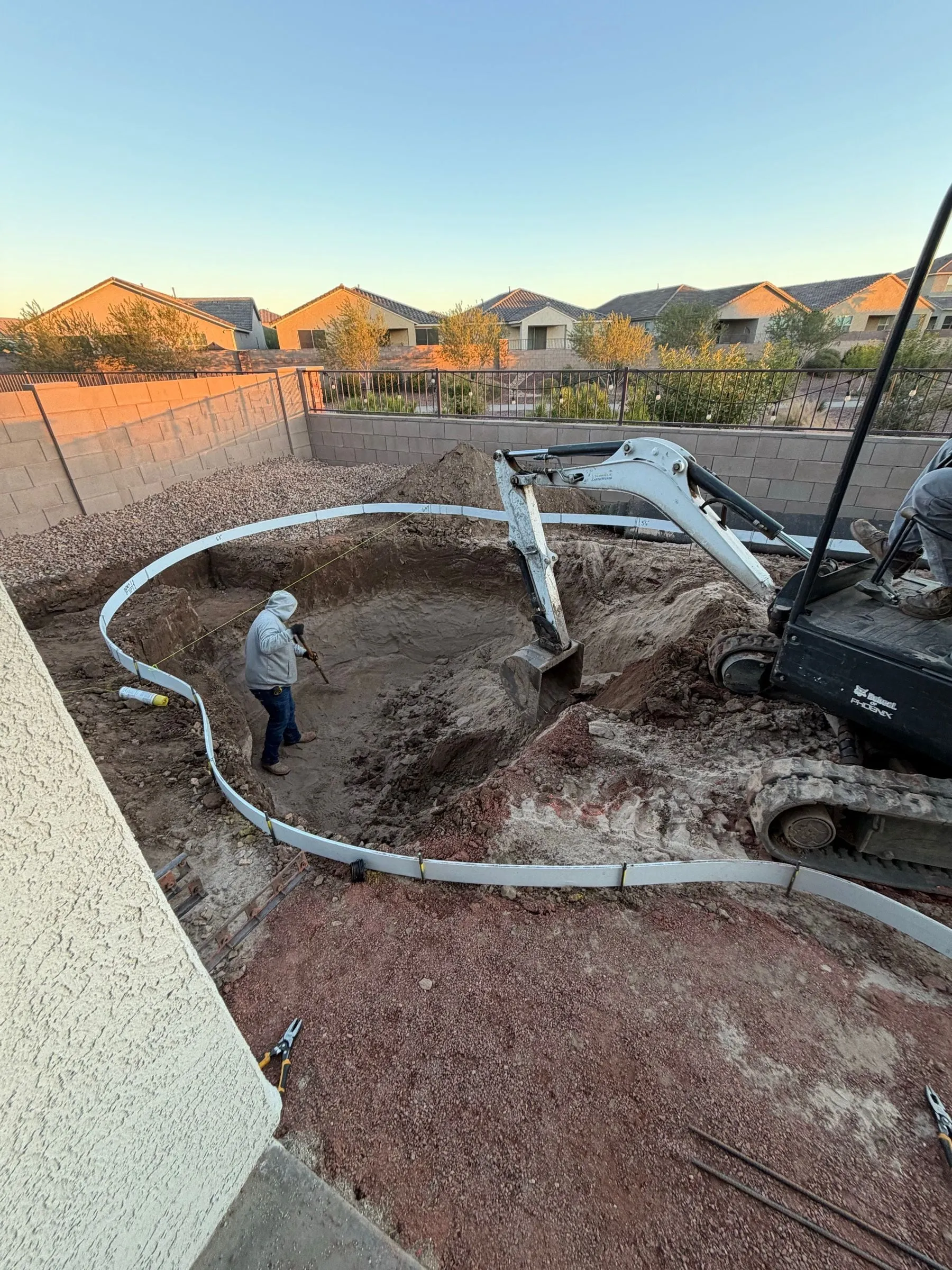 Mini excavator digging a freeform pool inside curved forms in the late-day light.
