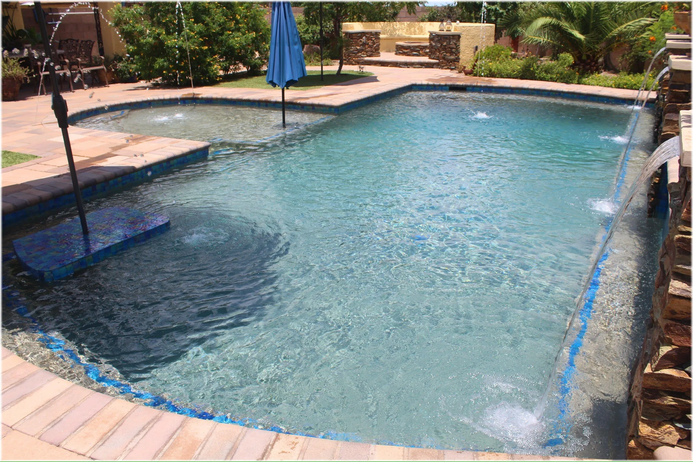 Freeform pool with glass tile waterline, tanning ledge umbrella, laminar jets and stone waterfall wall.
