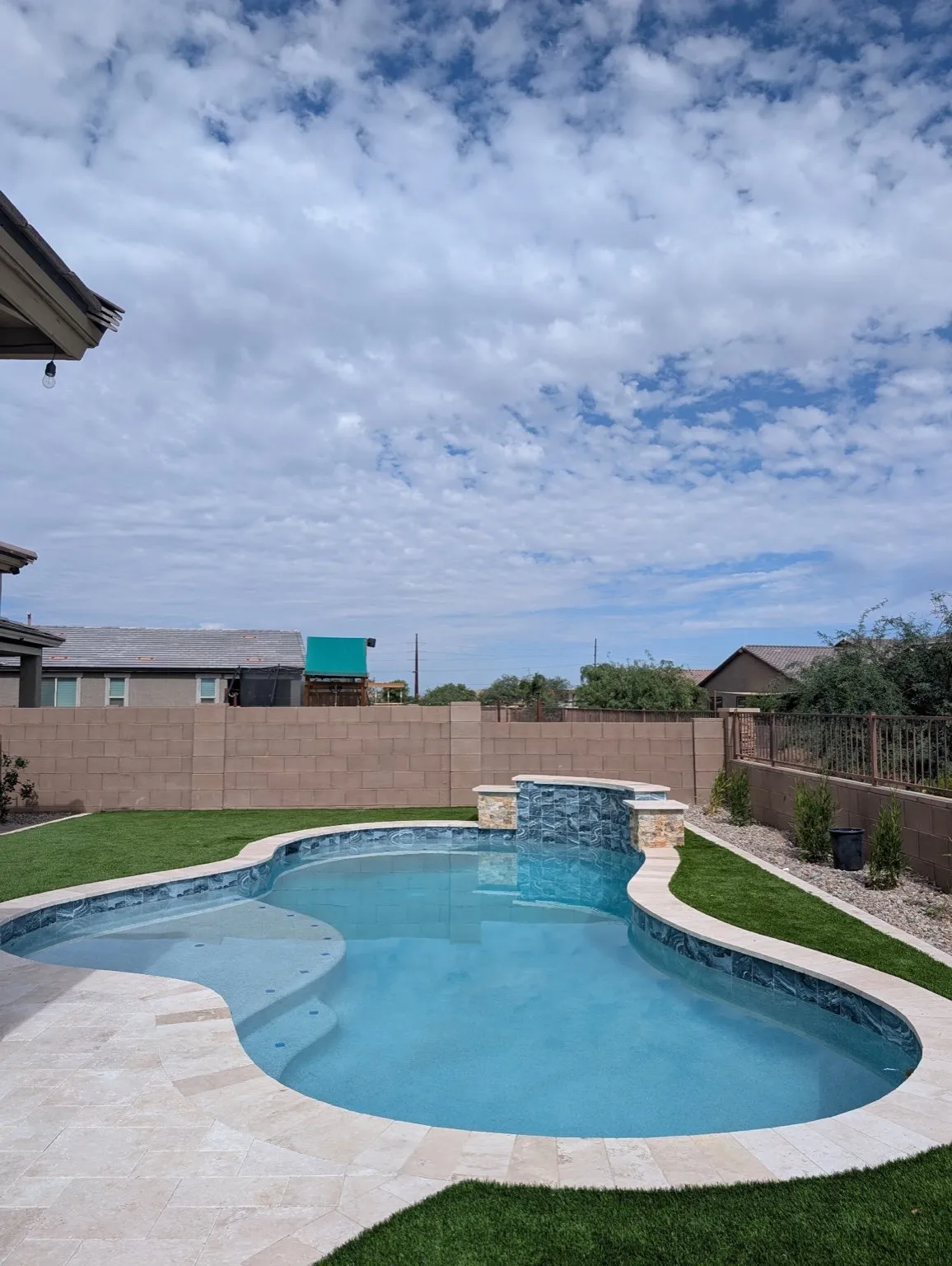 Freeform pool with a raised spillway wall, travertine deck and artificial turf under a cloudy Arizona sky.