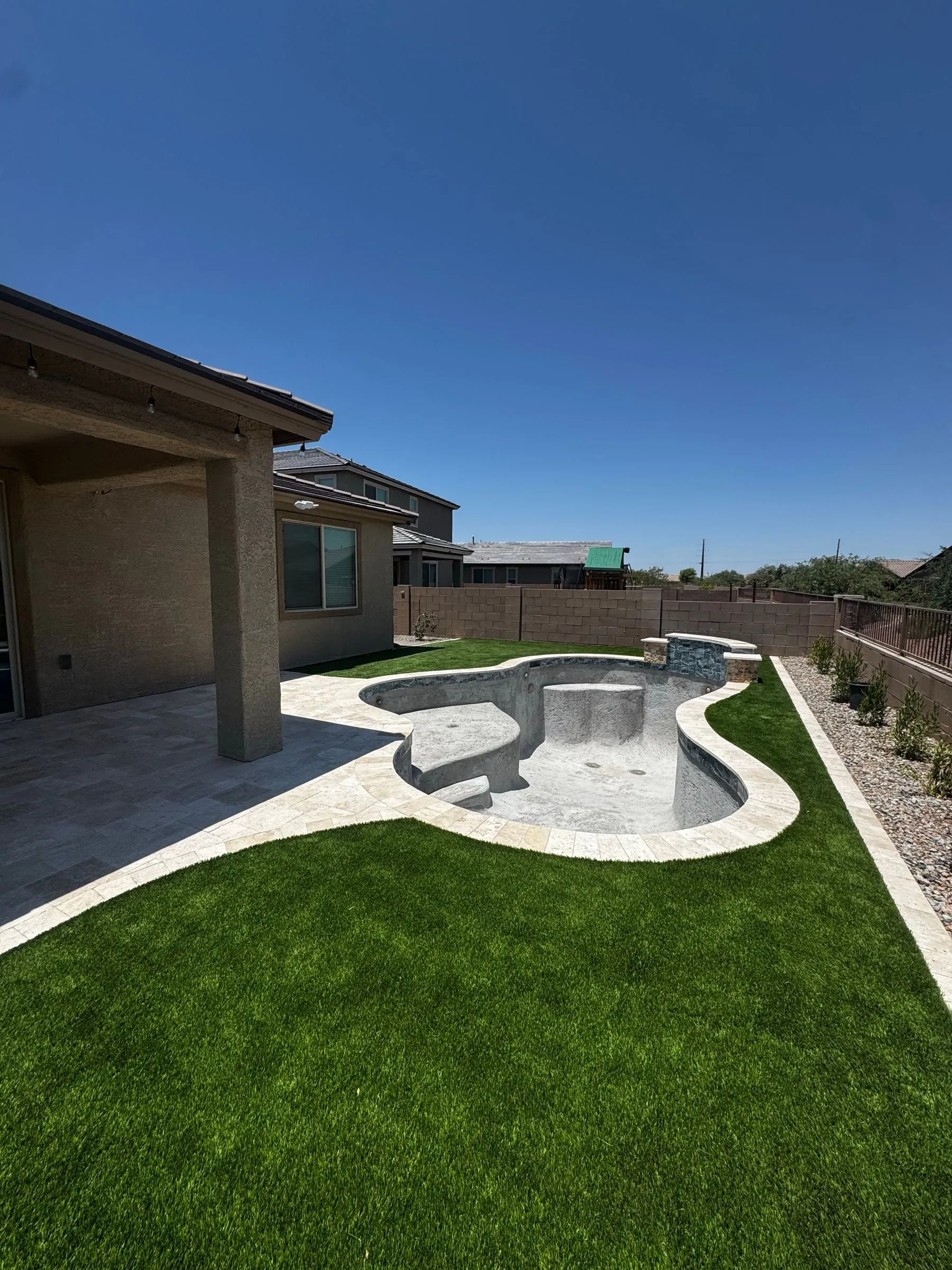 Freeform pool with travertine coping and deck curving through new artificial turf near a covered patio.