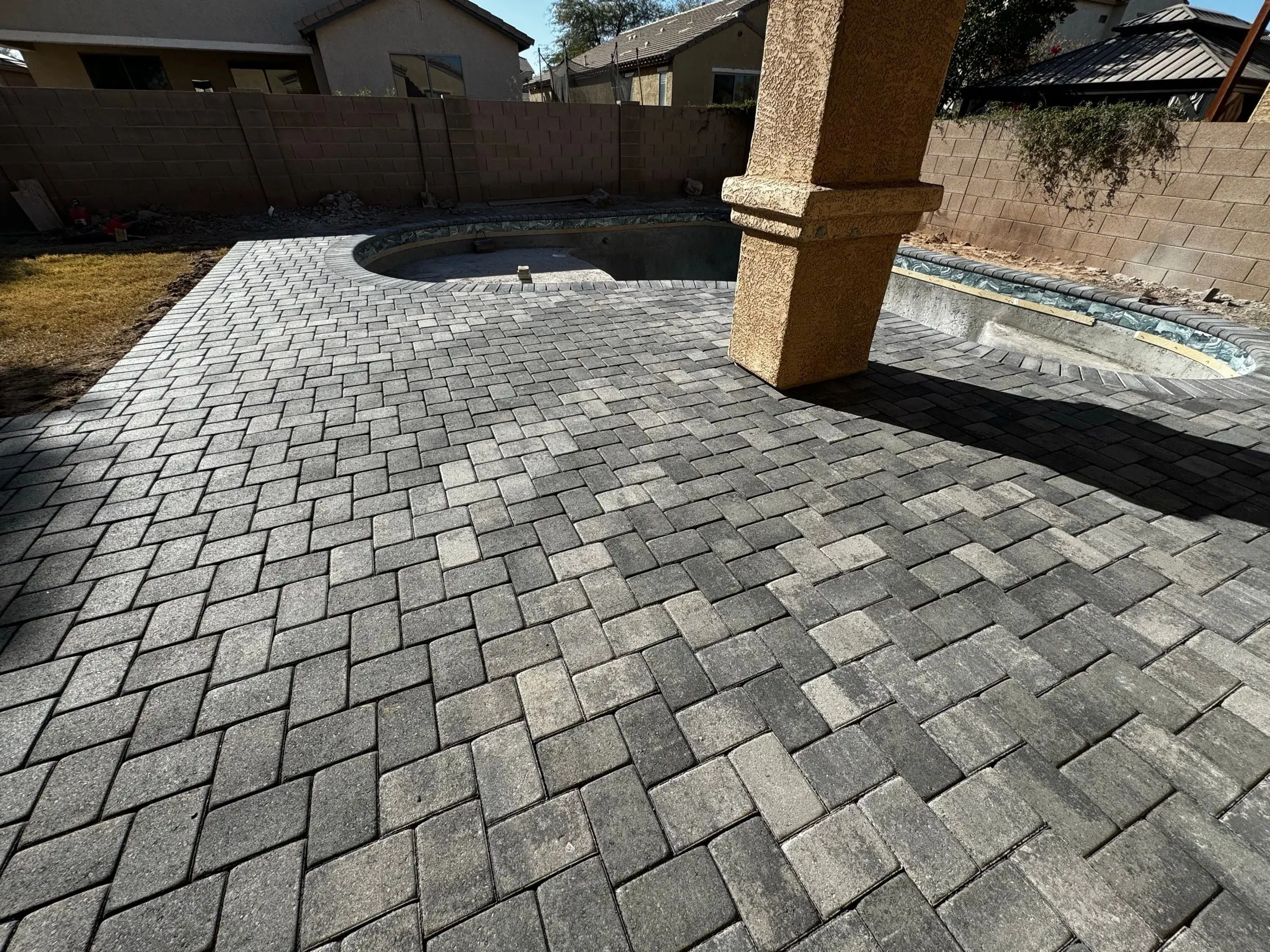 Gray herringbone paver patio installed under a covered porch beside a freeform pool shell.