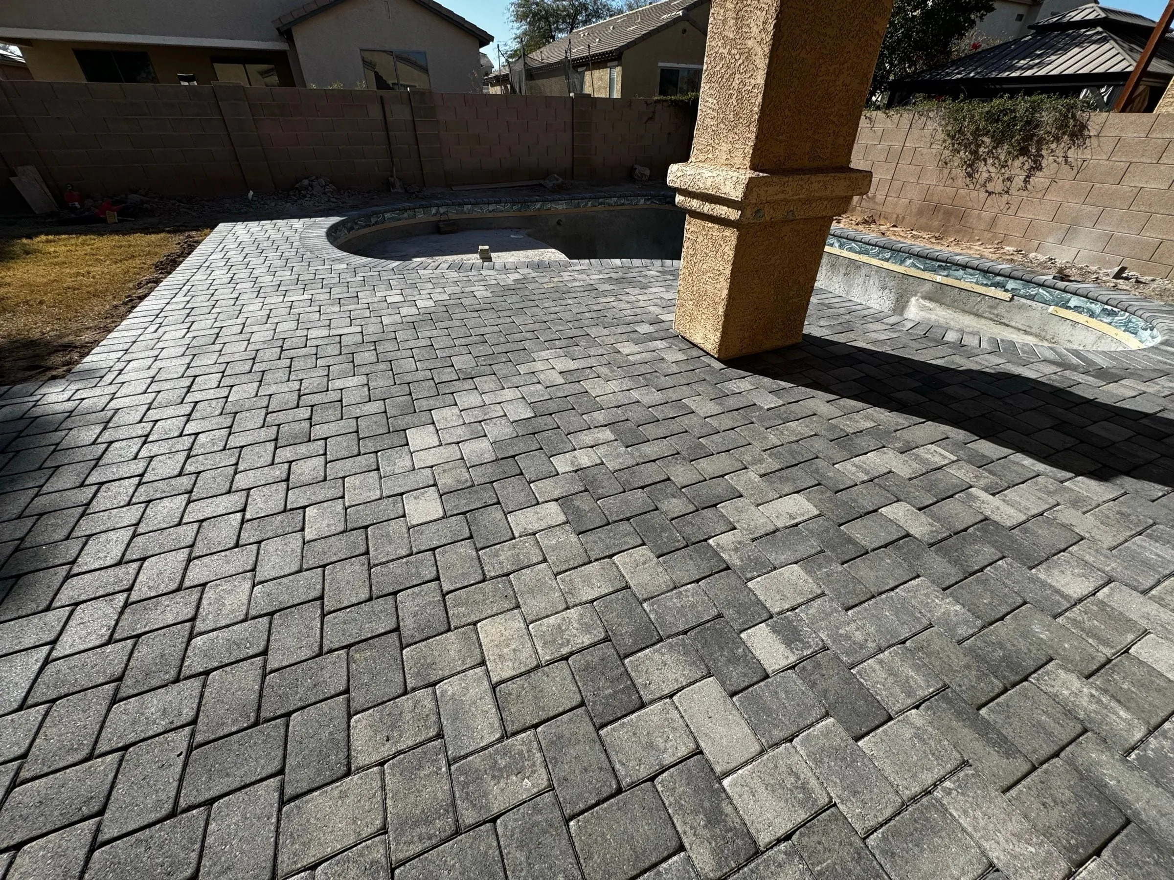 Gray herringbone paver patio installed under a covered porch beside a freeform pool shell.