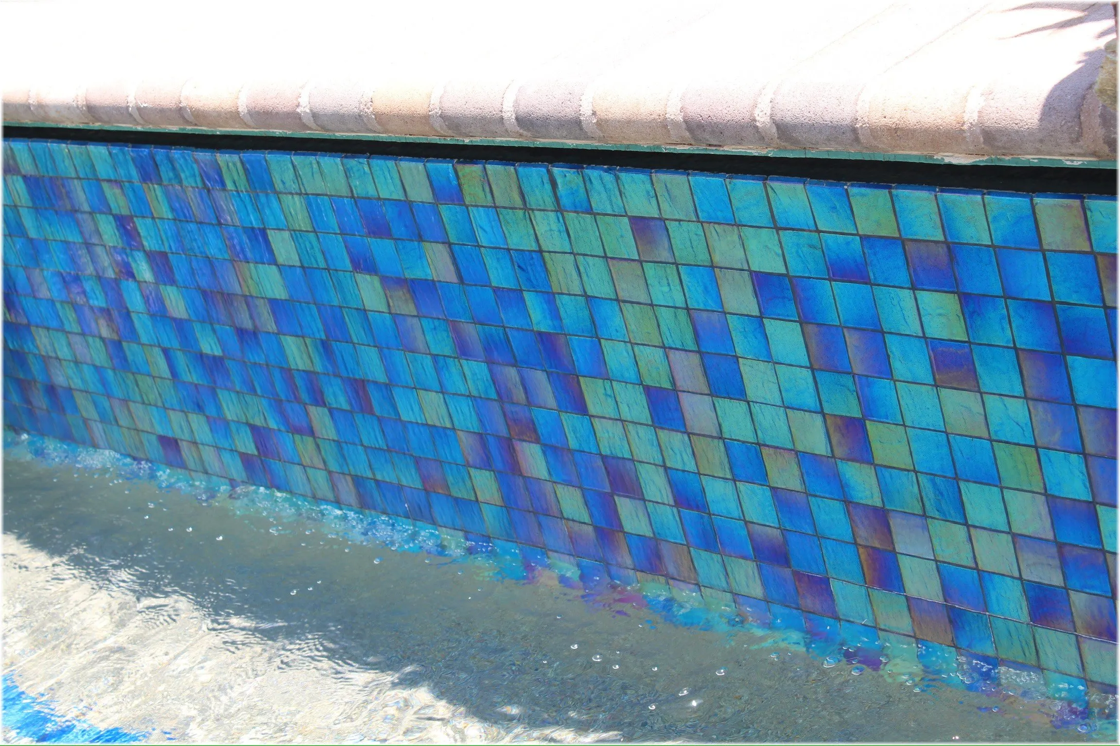 Close-up of iridescent blue glass tile along a pool waterline below the coping.