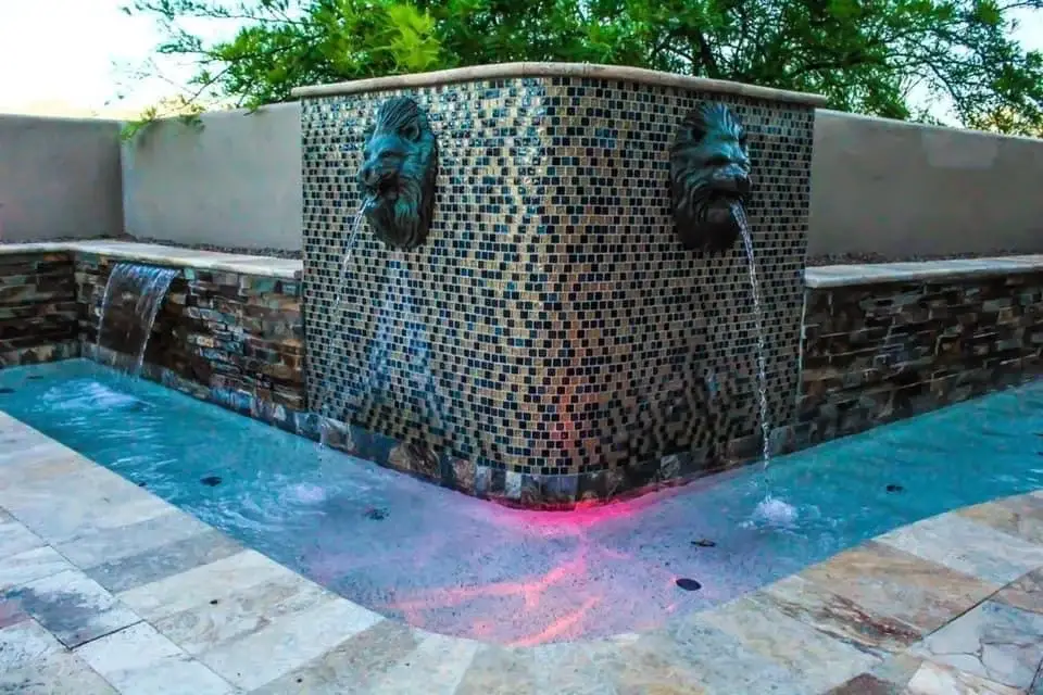Glass mosaic tile wall with bronze lion head spouts and a sheer descent above travertine.