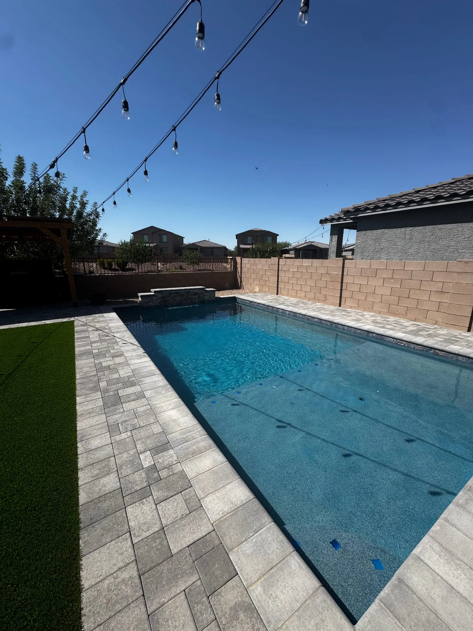 Long rectangular pool under string lights with a gray paver deck and turf.