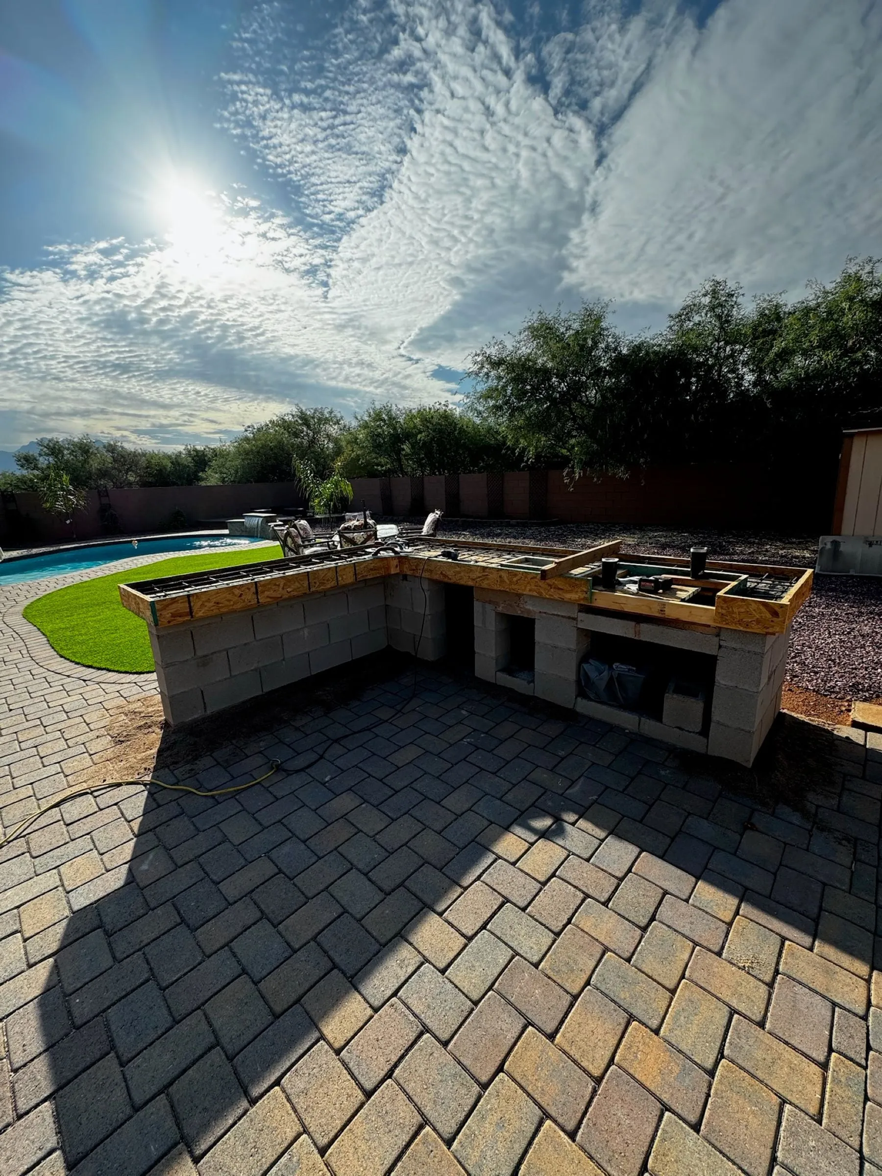 Block outdoor kitchen island under construction on a new paver patio beside a pool.
