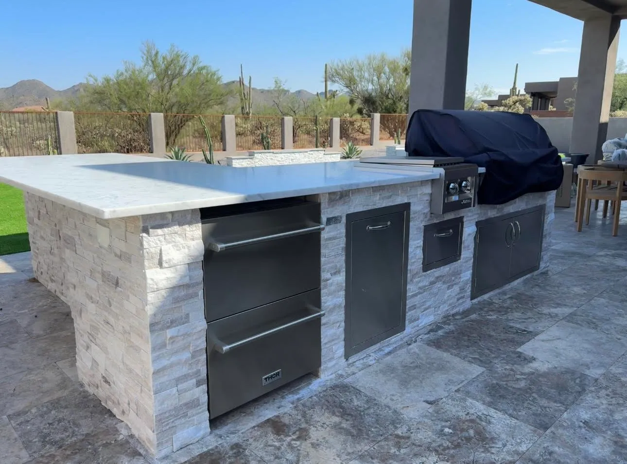Outdoor kitchen island with stacked stone base, white stone countertop and stainless appliances on travertine.