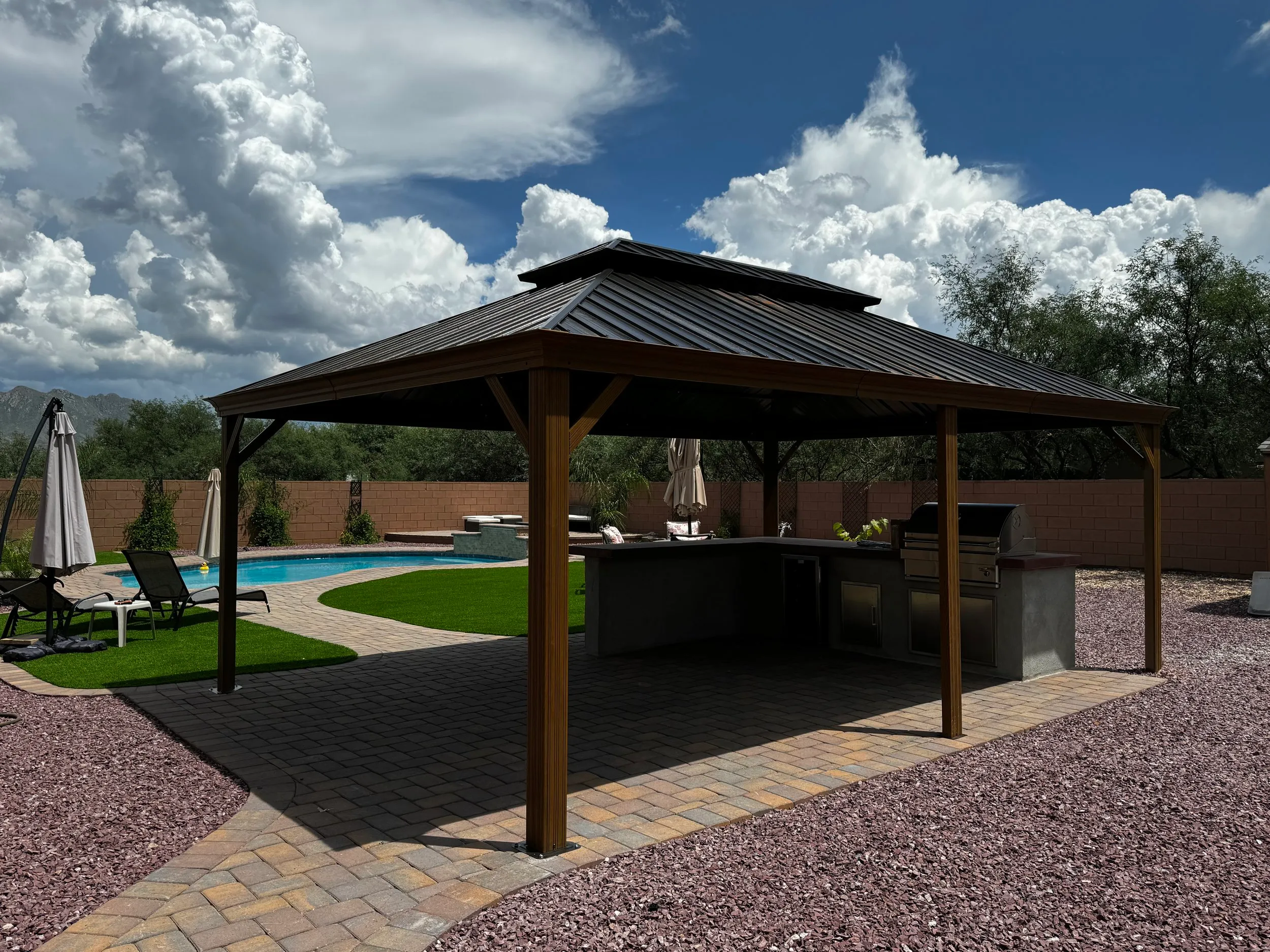 Metal-roof pergola over an outdoor kitchen next to a pool and paver patio