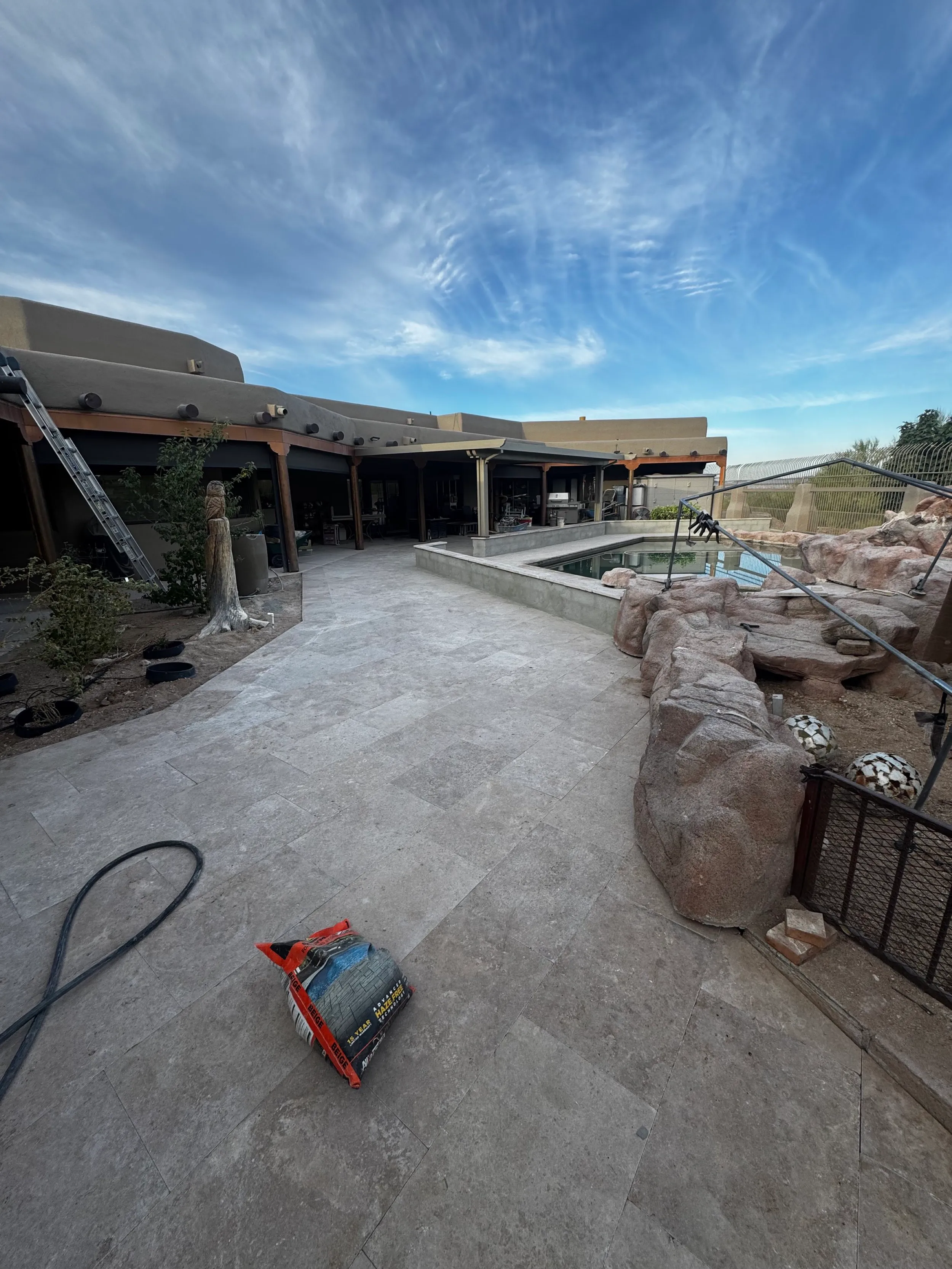 Large travertine patio and boulder water feature around a pool beside an adobe-style home