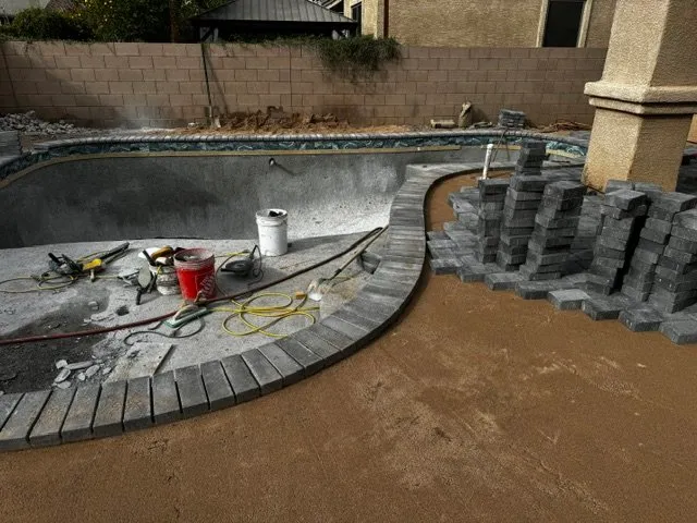 Paver coping being installed around a freeform pool with tools and stacked pavers nearby.