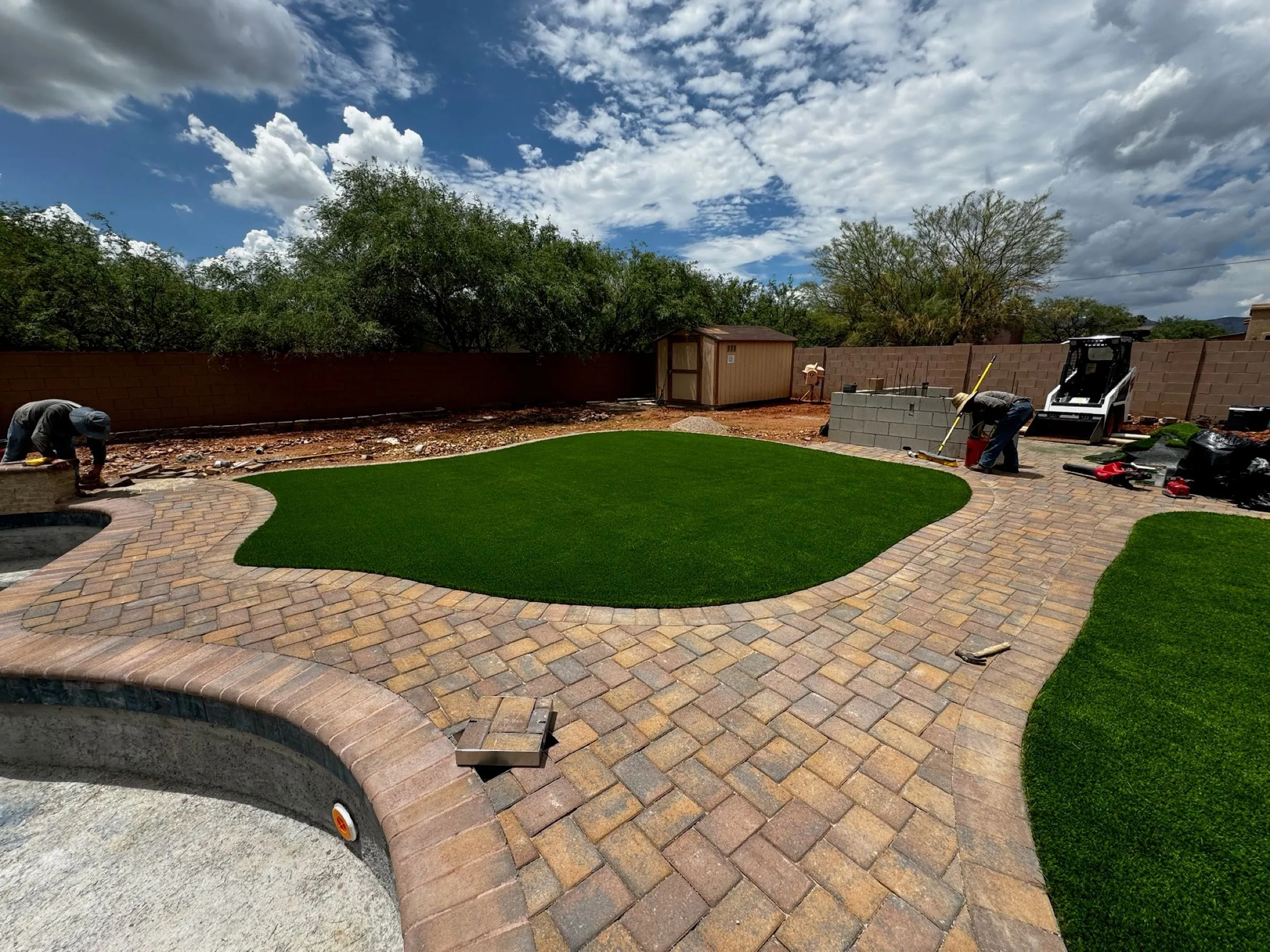 New brick-pattern paver deck and curved artificial turf beds being finished around a backyard pool.