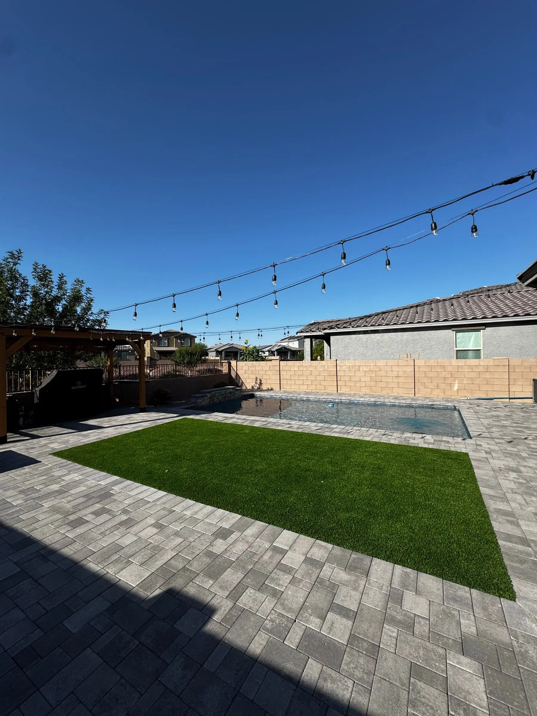 Gray paver deck with a turf panel and string lights beside a rectangular pool