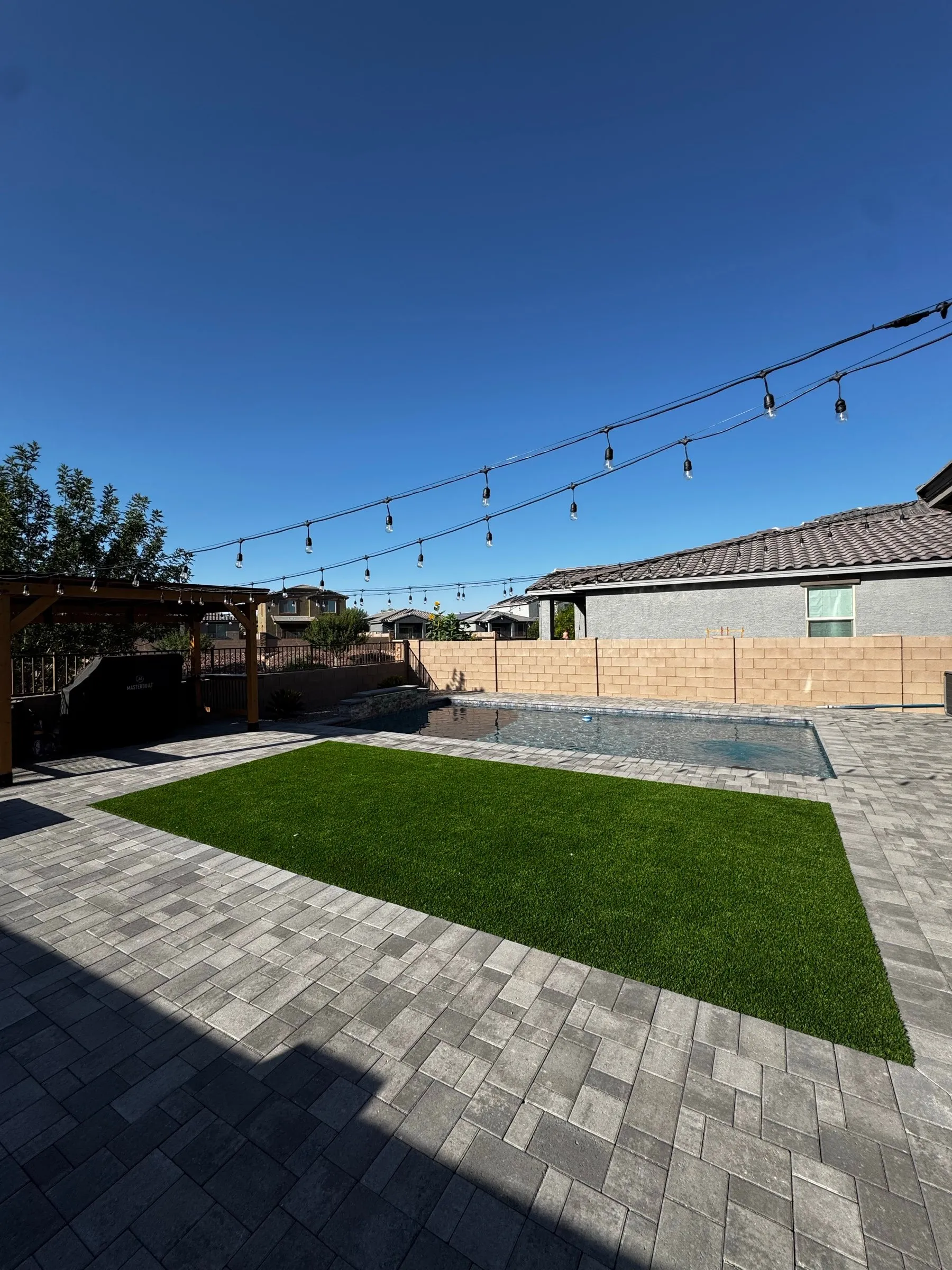 Gray paver deck with a turf panel and string lights beside a rectangular pool.