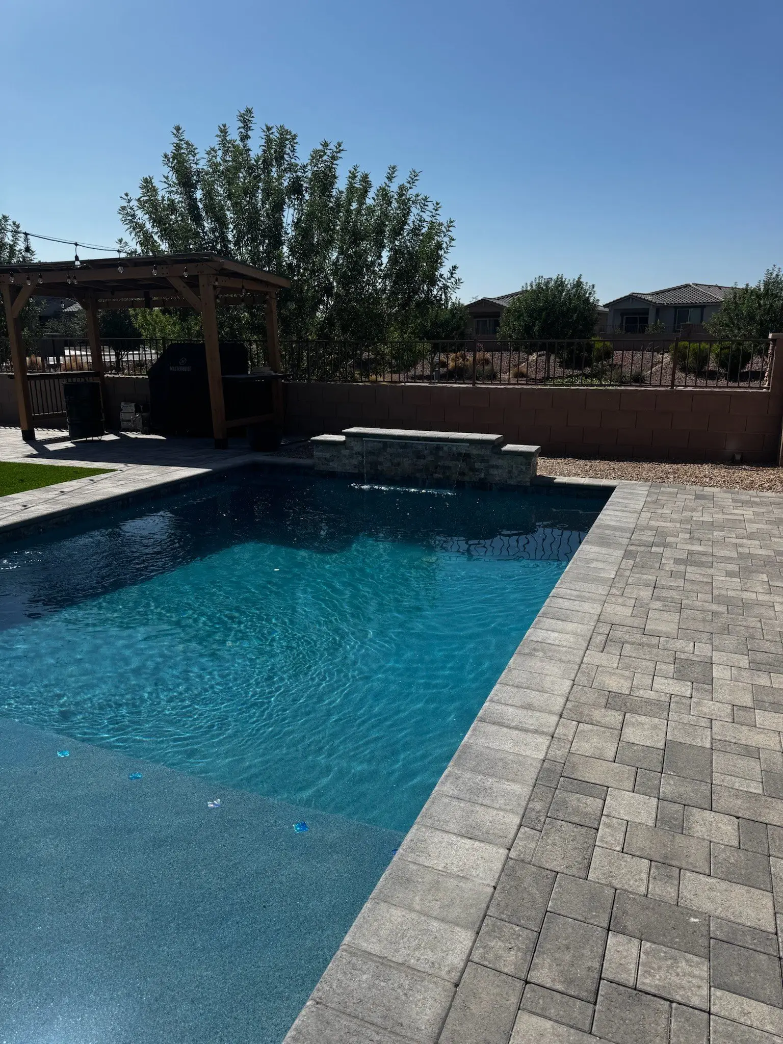 Rectangular pool with paver deck, raised spillway and a wood pergola over an outdoor grill.