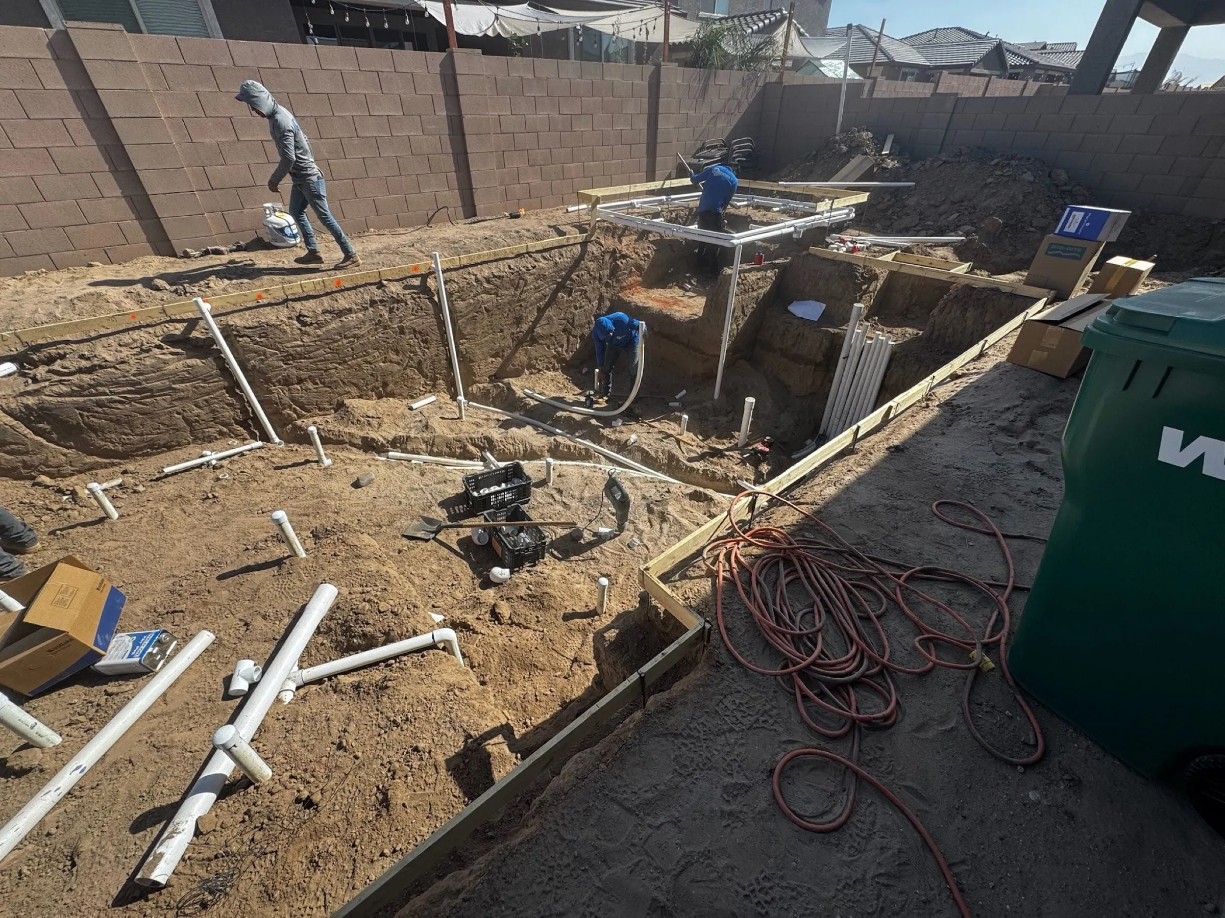 Crew members work in a rectangular pool dig as PVC plumbing lines are laid out.
