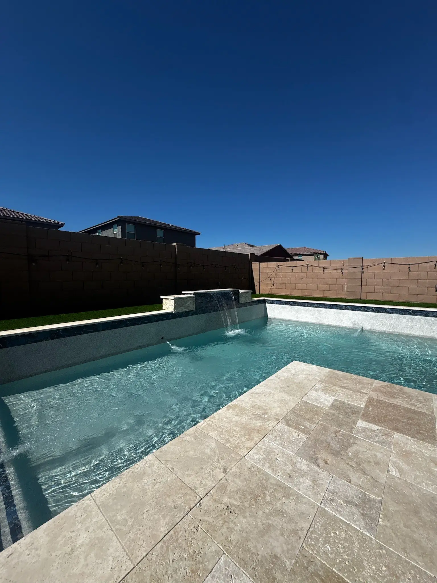 Pool with raised spillway, travertine deck, string lights and artificial turf on a sunny Arizona day.