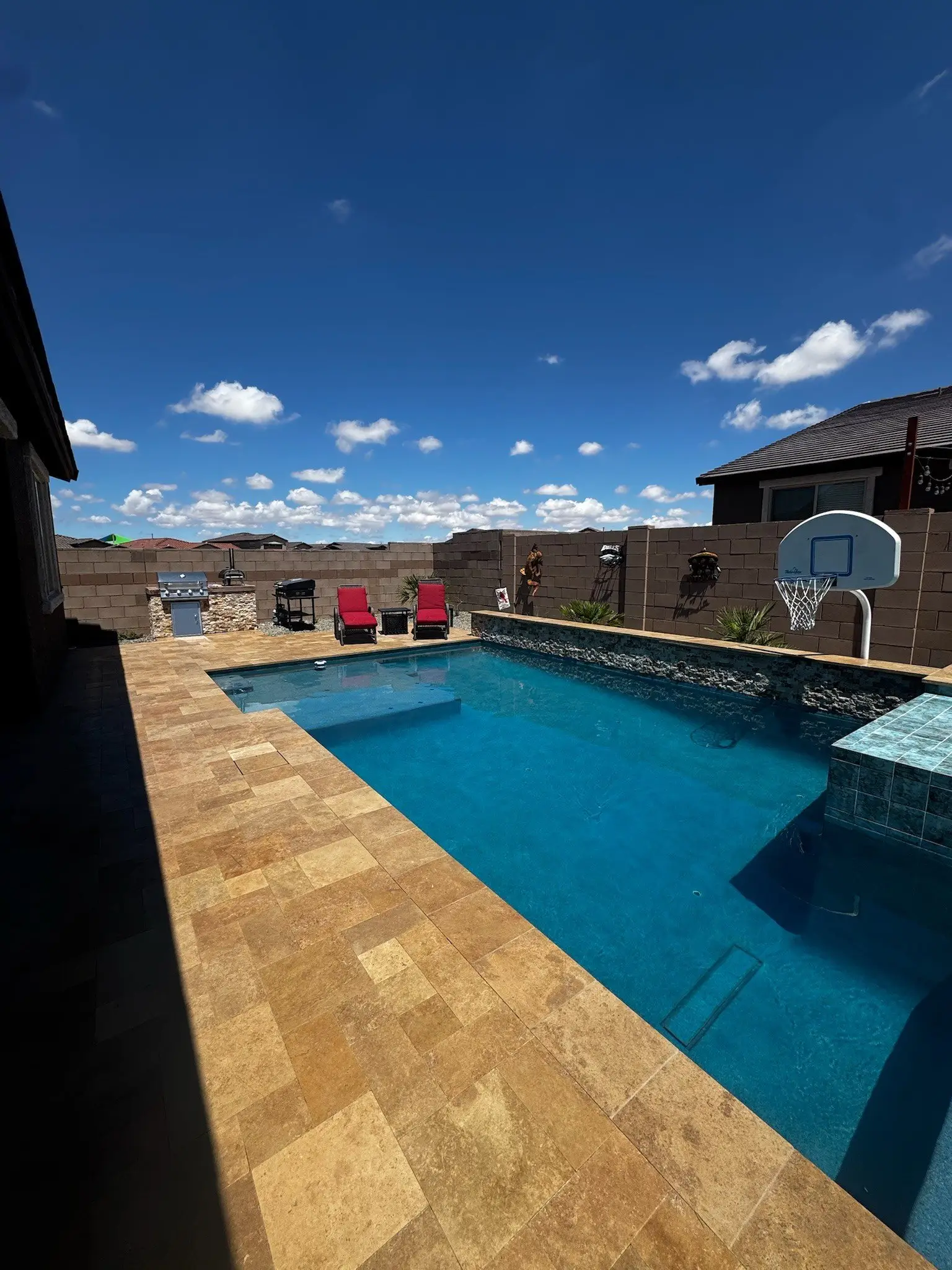 Wide view of a rectangular pool with tanning ledge, gold travertine deck and stacked stone raised wall.