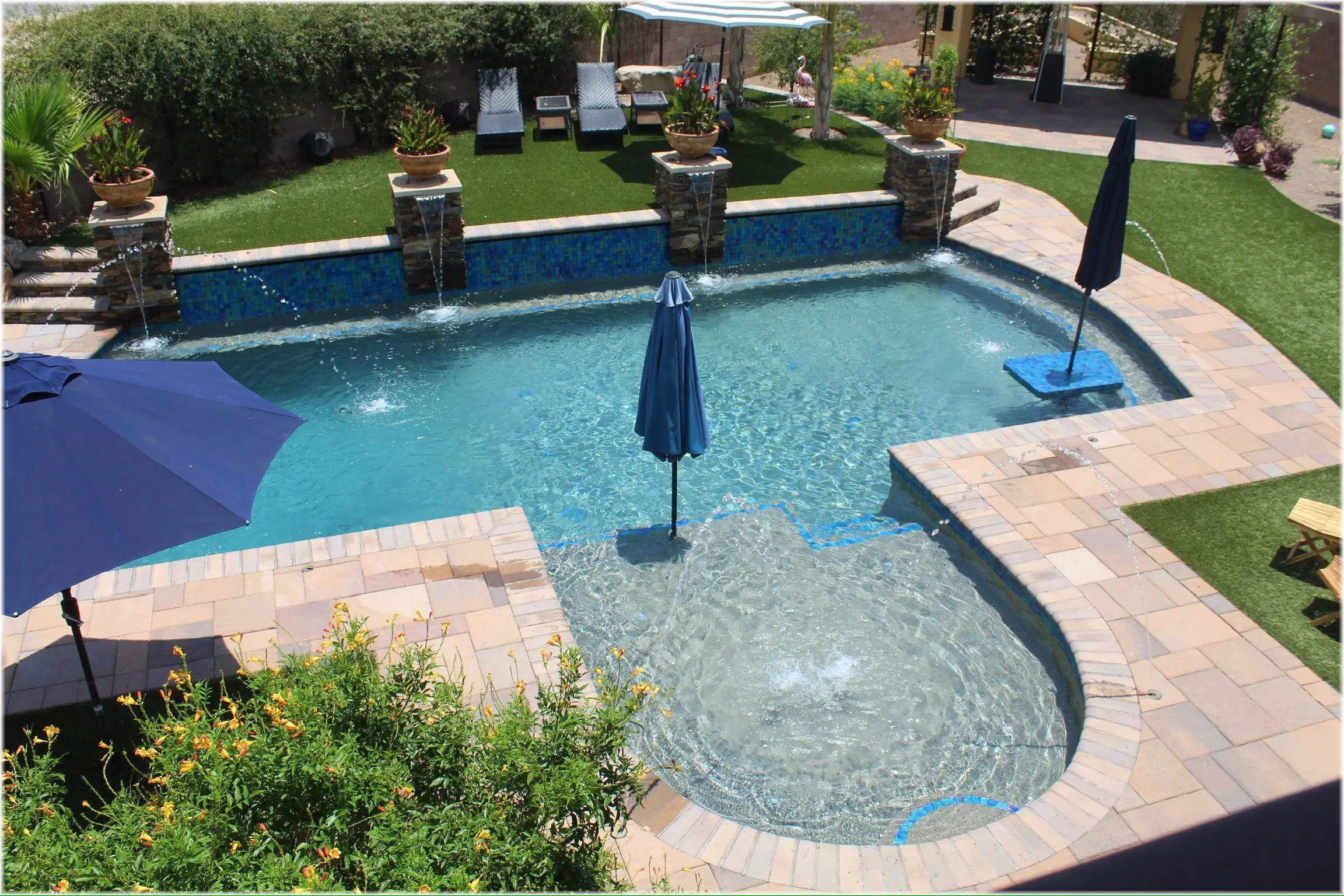 Pool with tanning ledge bubblers, shade umbrellas and stone pillar scuppers seen from above past flowering shrubs