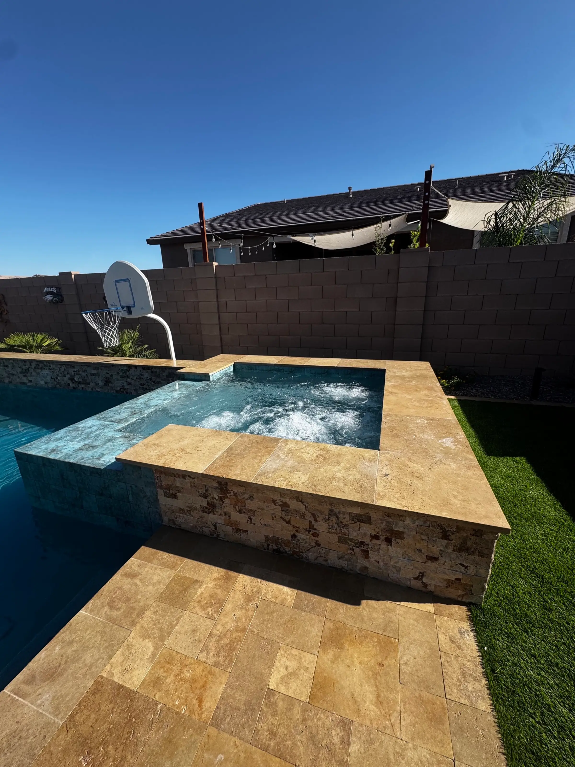 Raised spa with stacked stone base spilling into the pool, with travertine deck and artificial turf.
