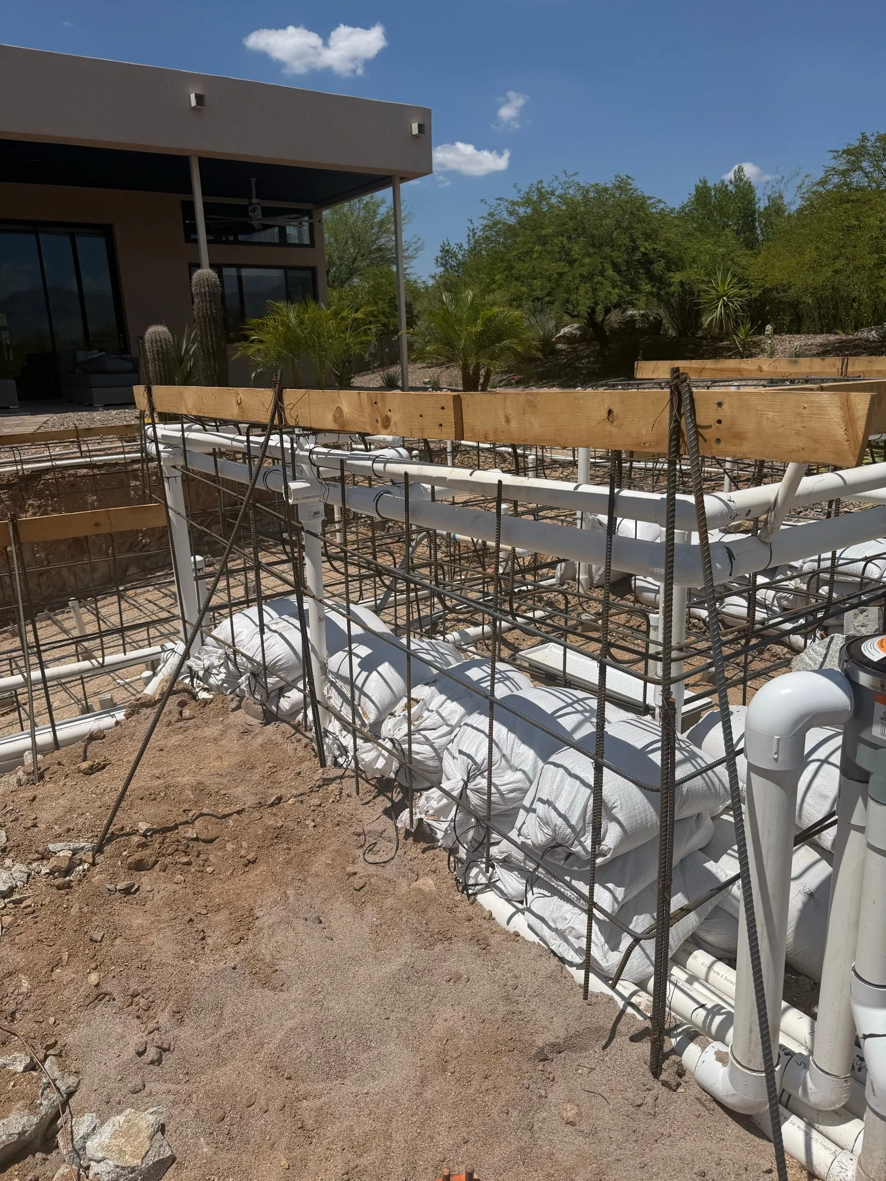 Rebar cage and PVC plumbing lines running along a raised pool wall braced with sandbags