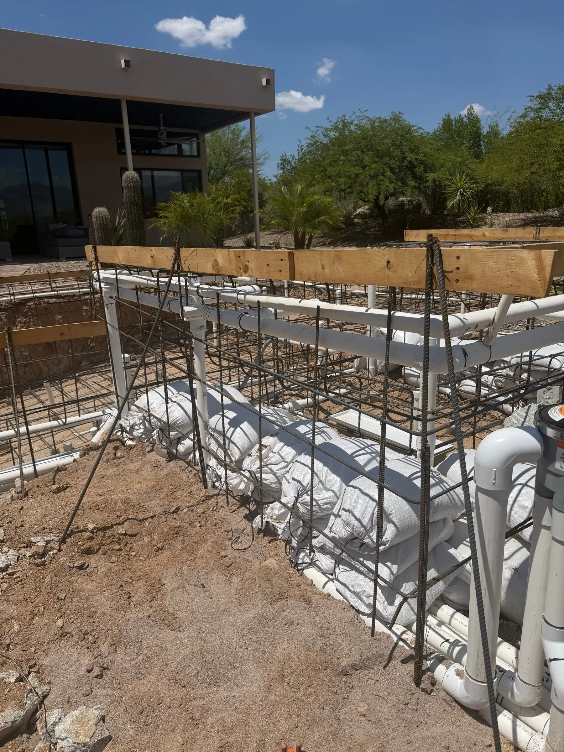 Rebar cage and PVC plumbing lines running along a raised pool wall braced with sandbags