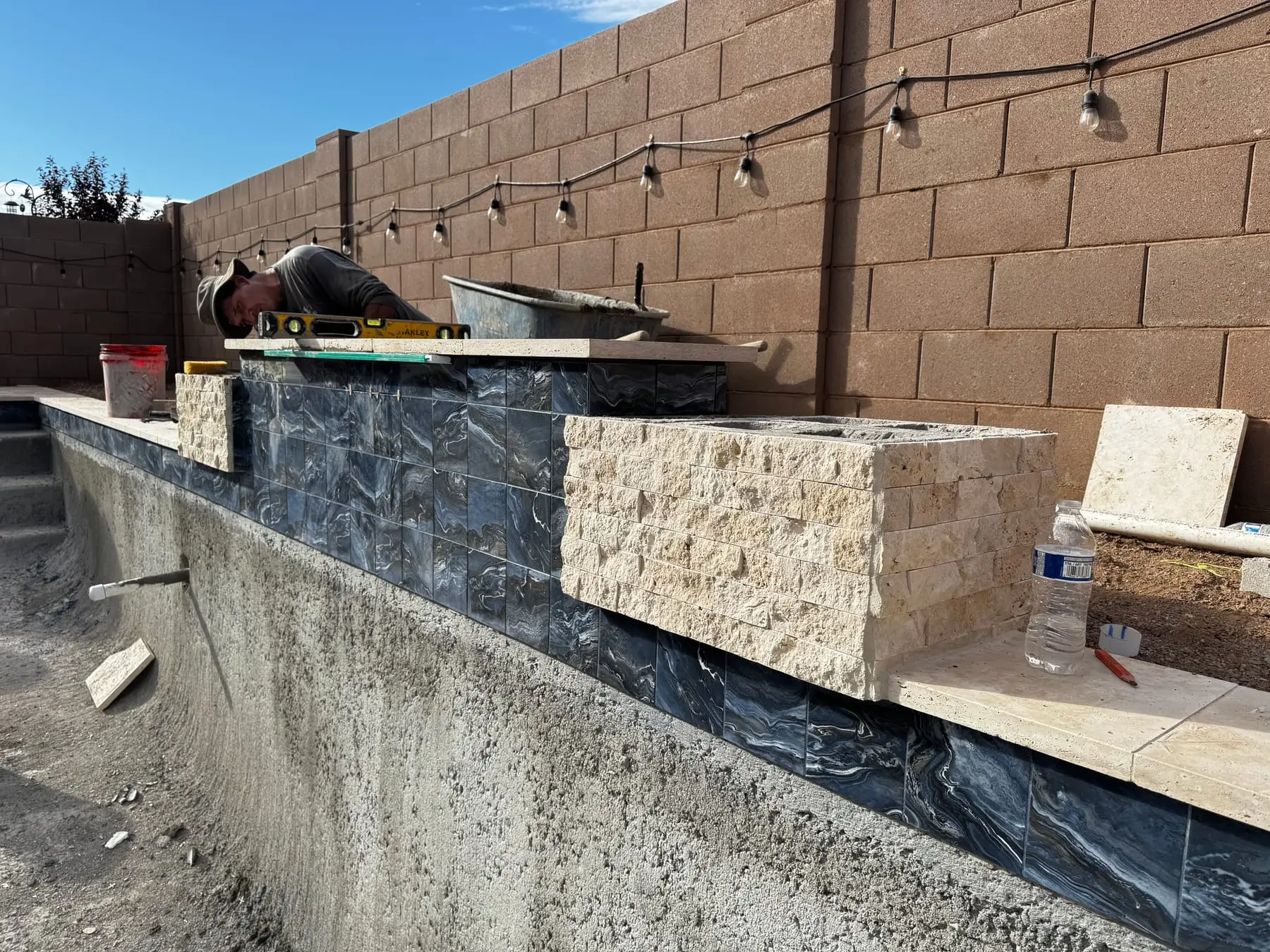 Raised wall getting stacked stone veneer and blue mosaic tile above a new pool shell.