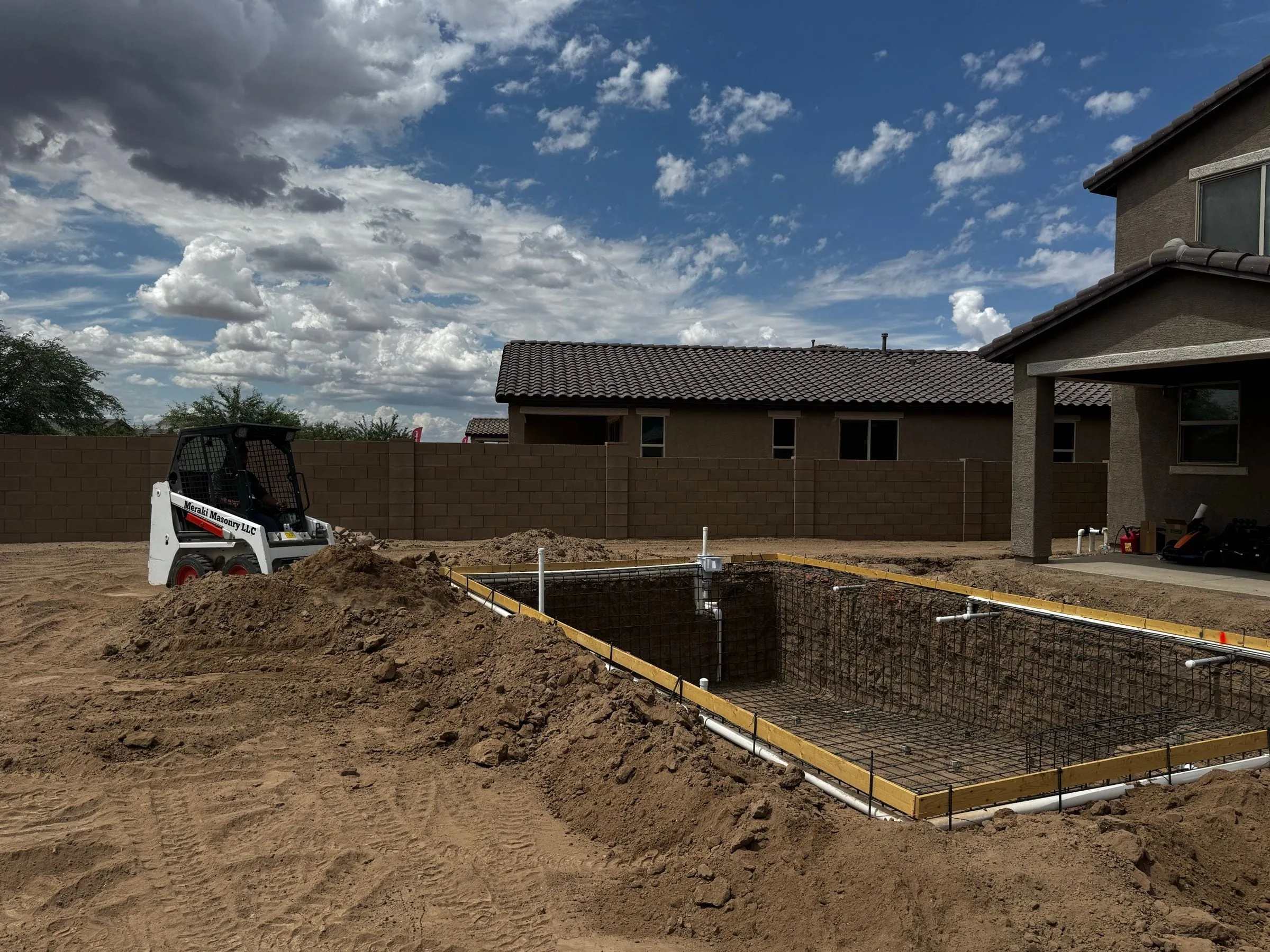 Rebar and plumbing set in a rectangular pool excavation, with a skid steer and dirt piles nearby.
