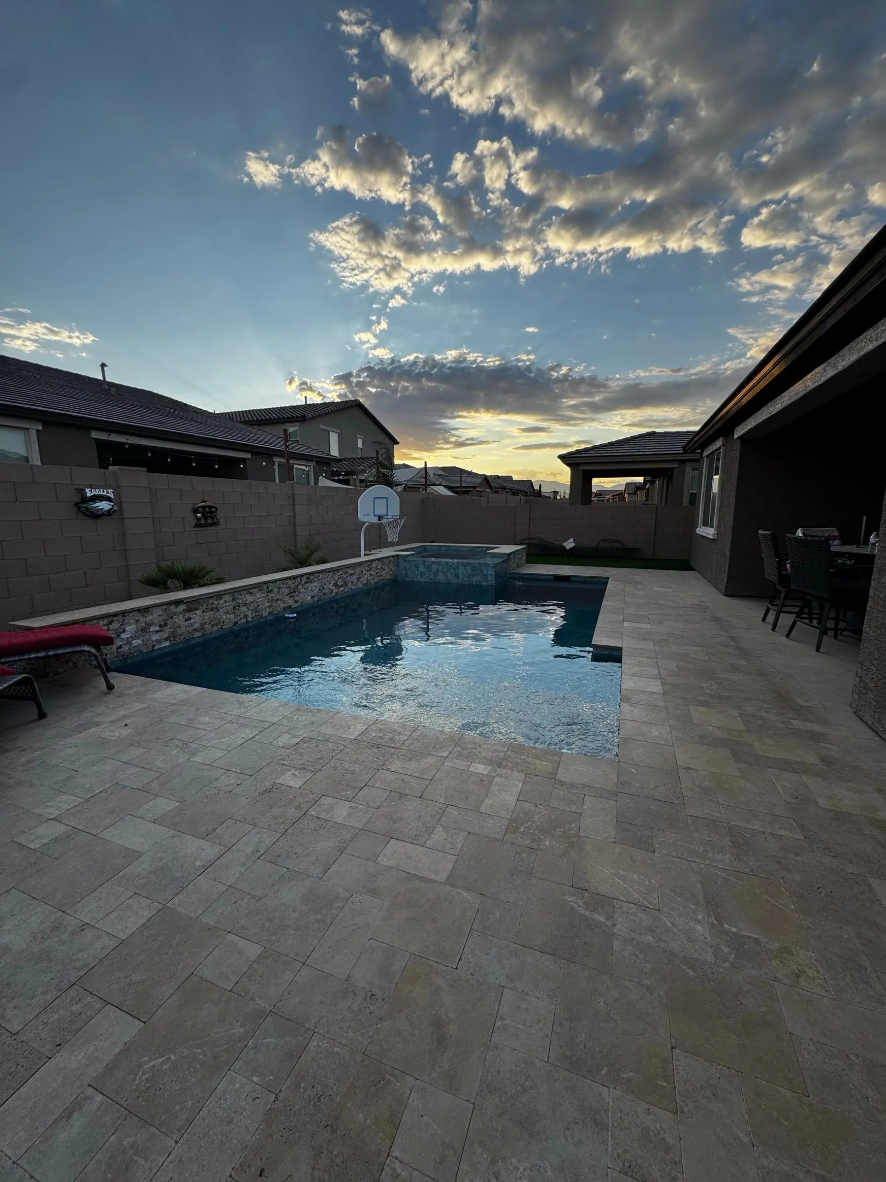 Rectangular pool with travertine deck and stacked stone raised wall under sunset clouds, Marana, AZ.
