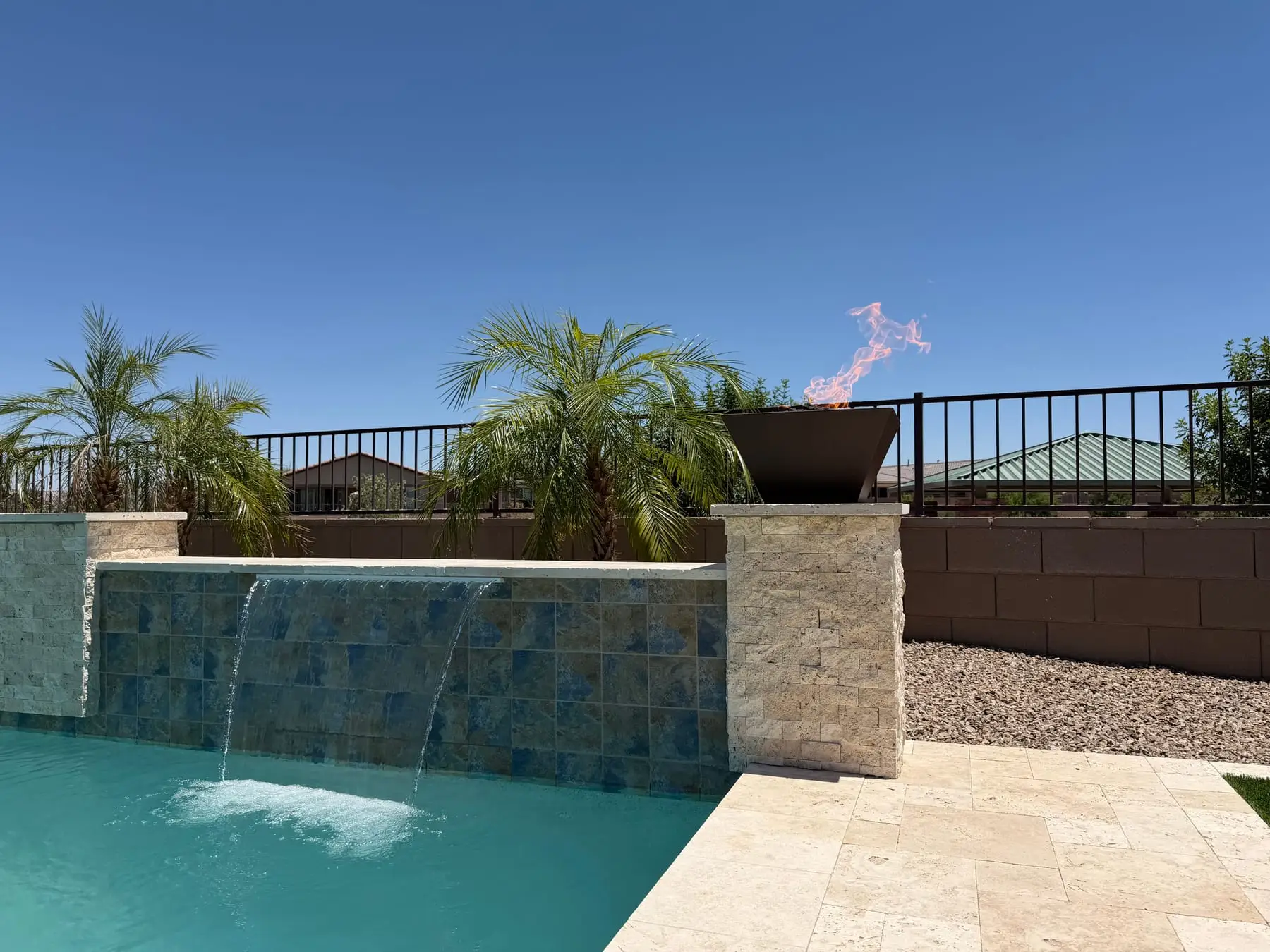 Sheer descent spilling from a glass-tile raised wall between two lit fire bowls on travertine pillars.