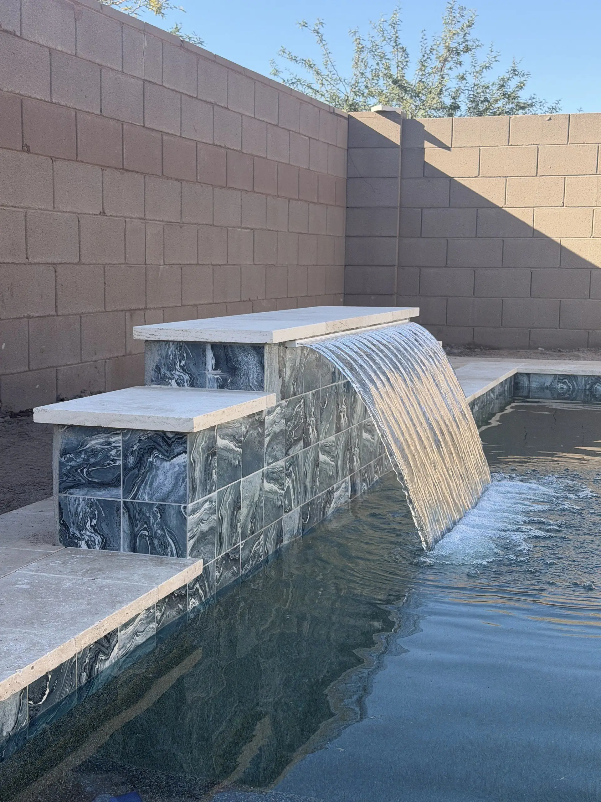 Close-up of a sheer descent flowing from a marble-look tile raised wall with travertine coping.