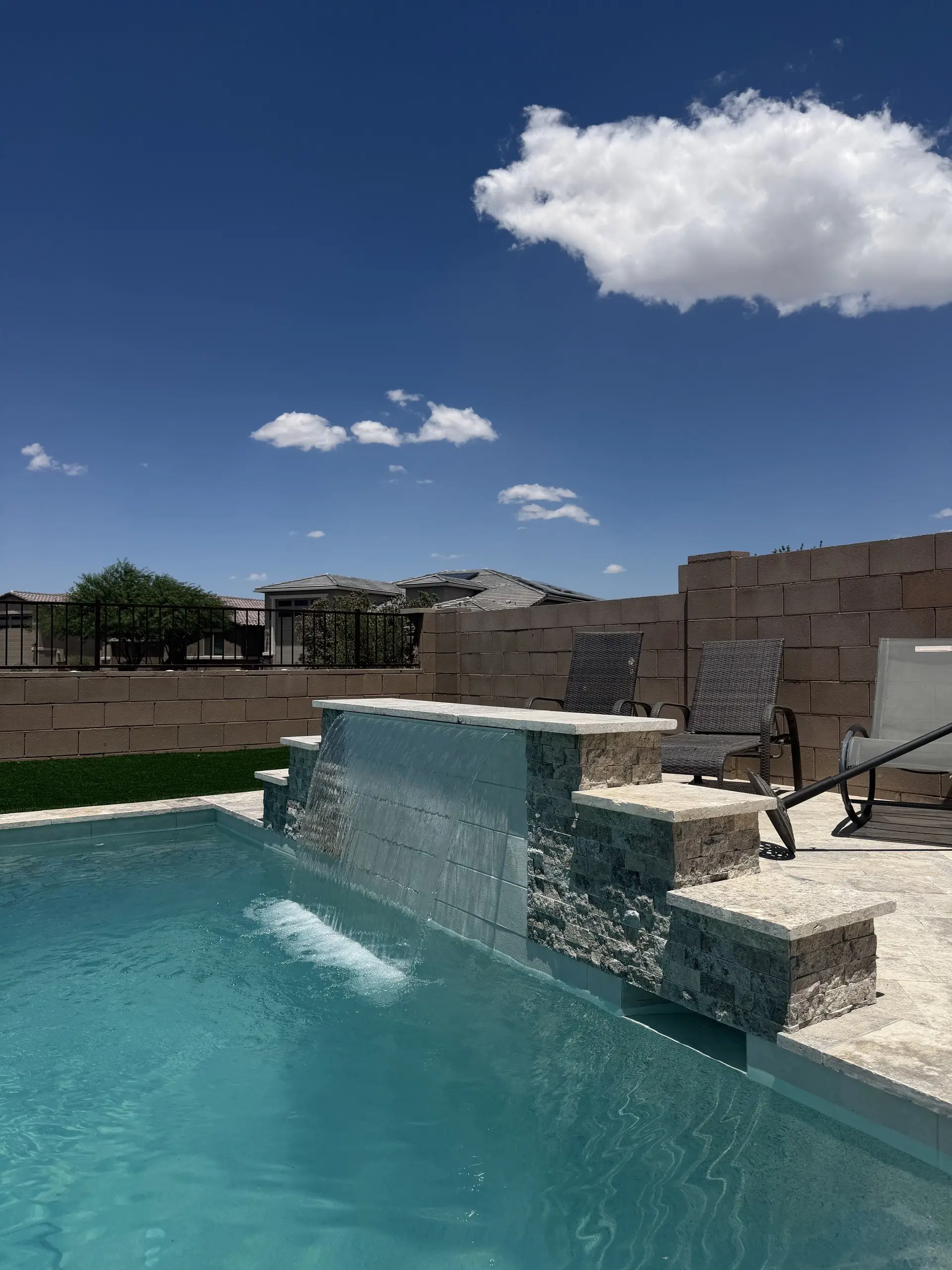 Sheer descent pouring from a stacked stone raised wall beside a travertine deck and lounge chairs.