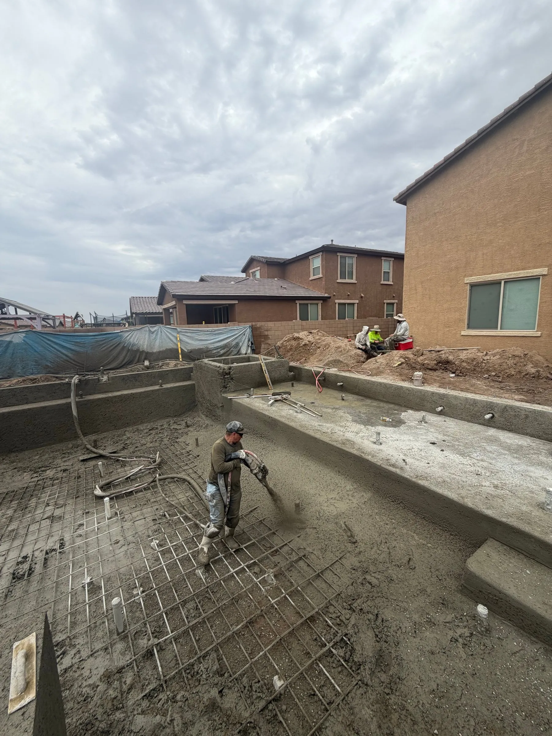 Nozzleman spraying shotcrete over the rebar floor of a rectangular pool under cloudy skies in Marana.