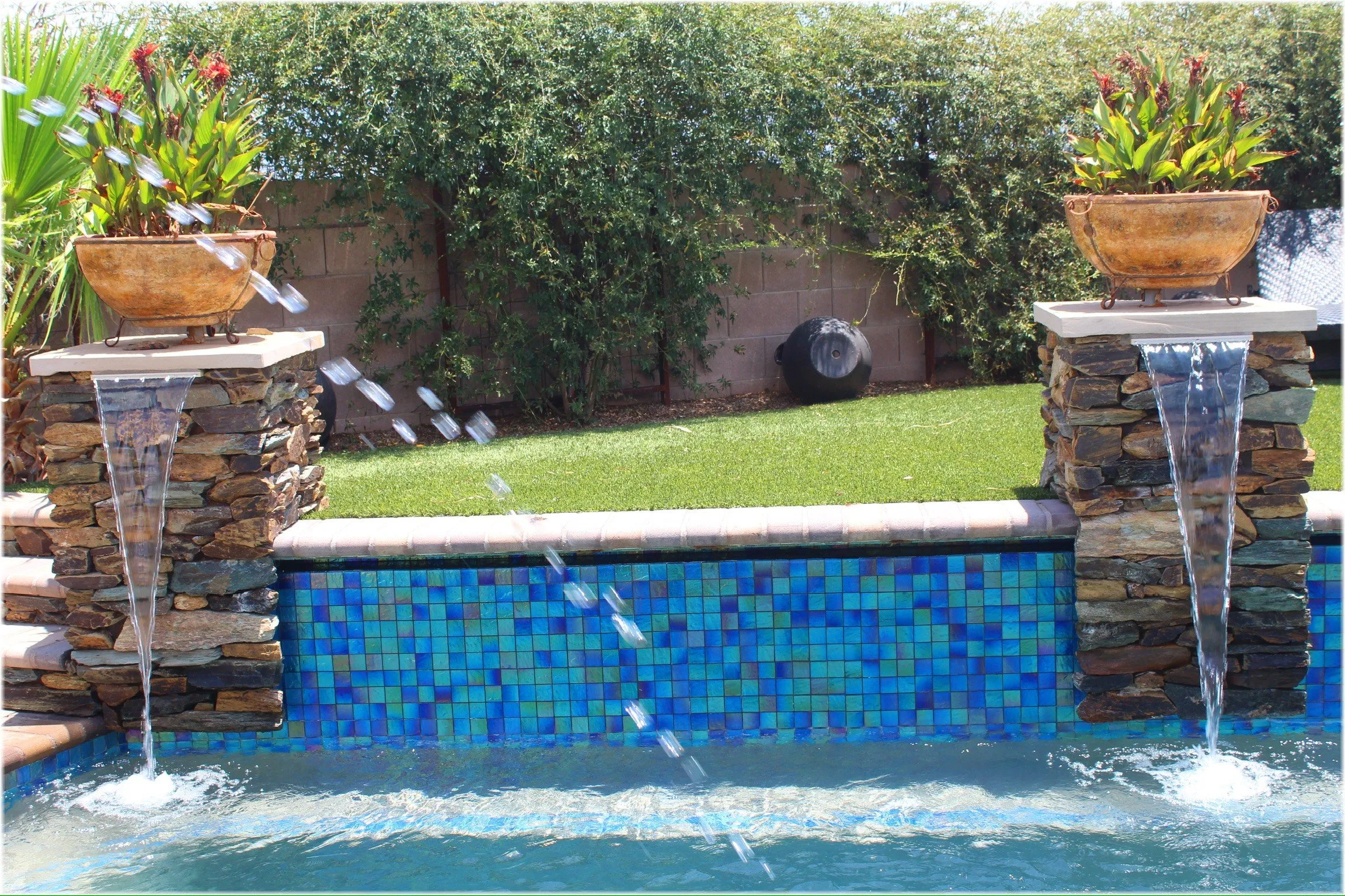 Stacked stone pillars with sheer descents and planters above a blue glass tile pool wall.