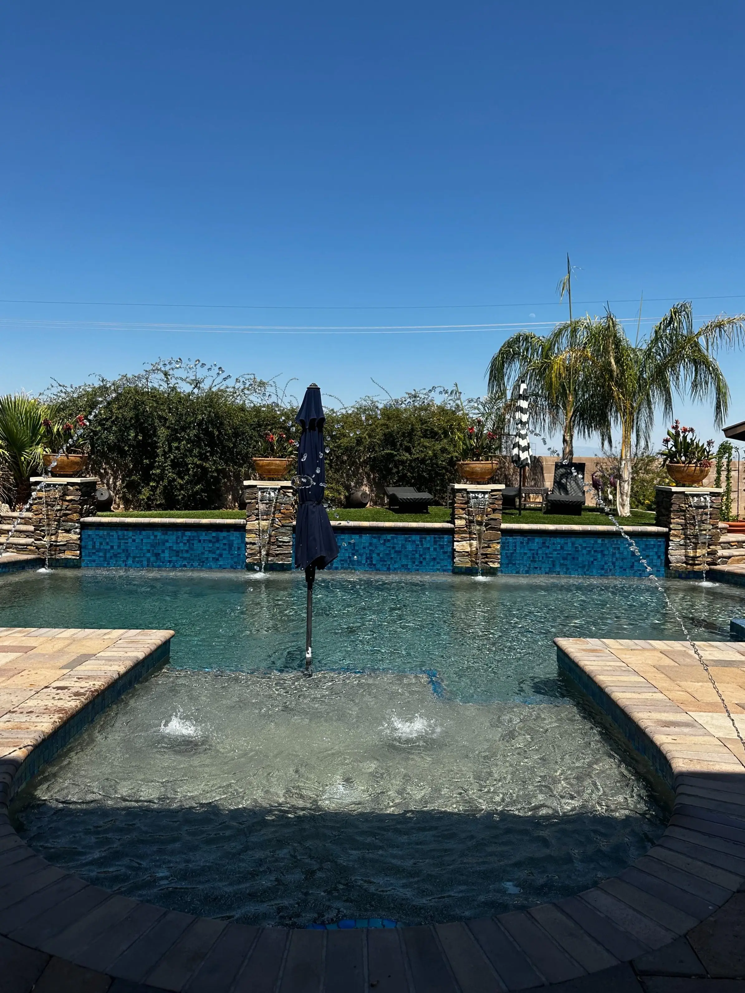 Tanning ledge bubblers and umbrella facing a blue glass tile wall with stacked stone scuppers.
