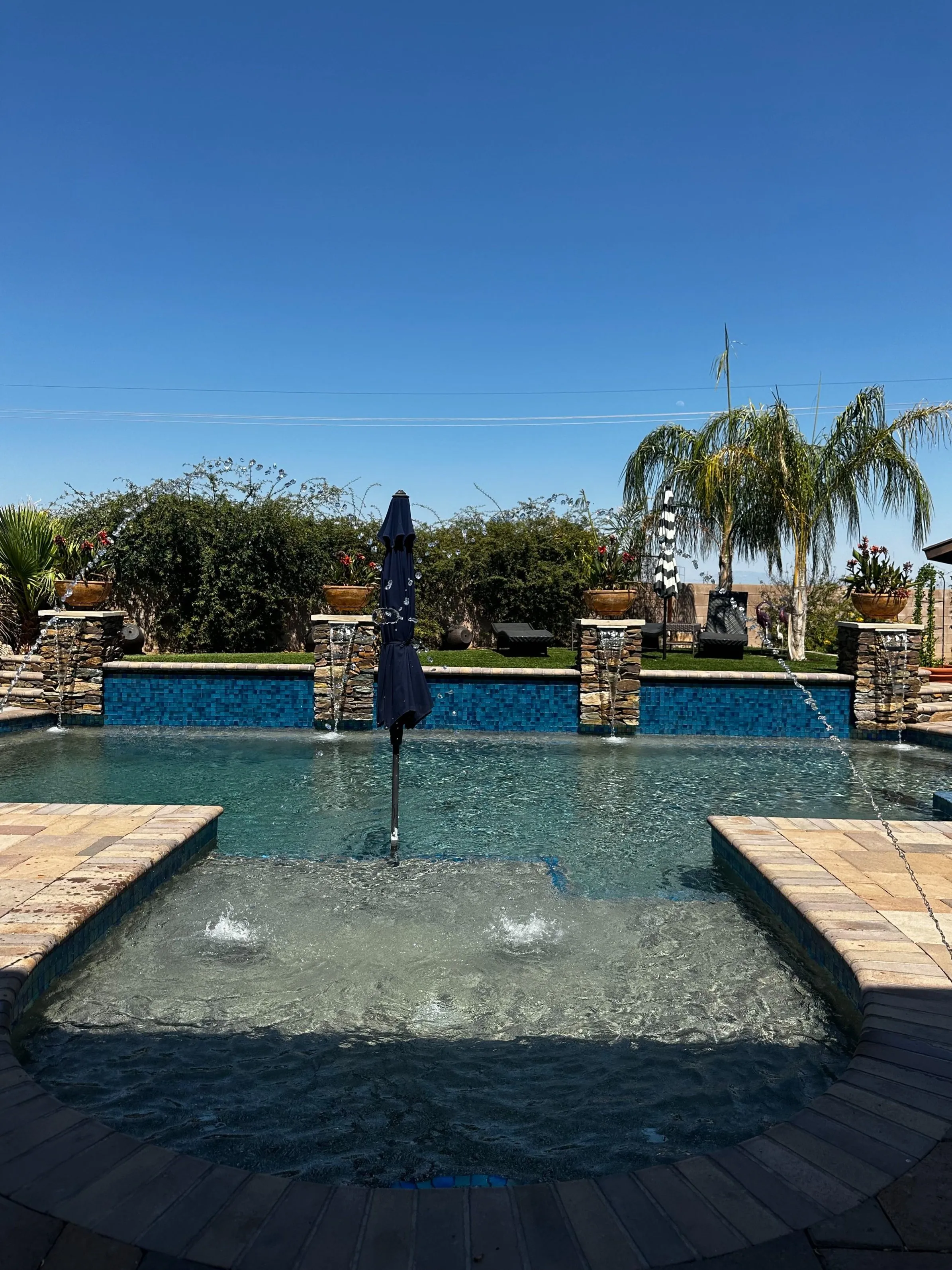 Tanning ledge bubblers and umbrella facing a blue glass tile wall with stacked stone scuppers.
