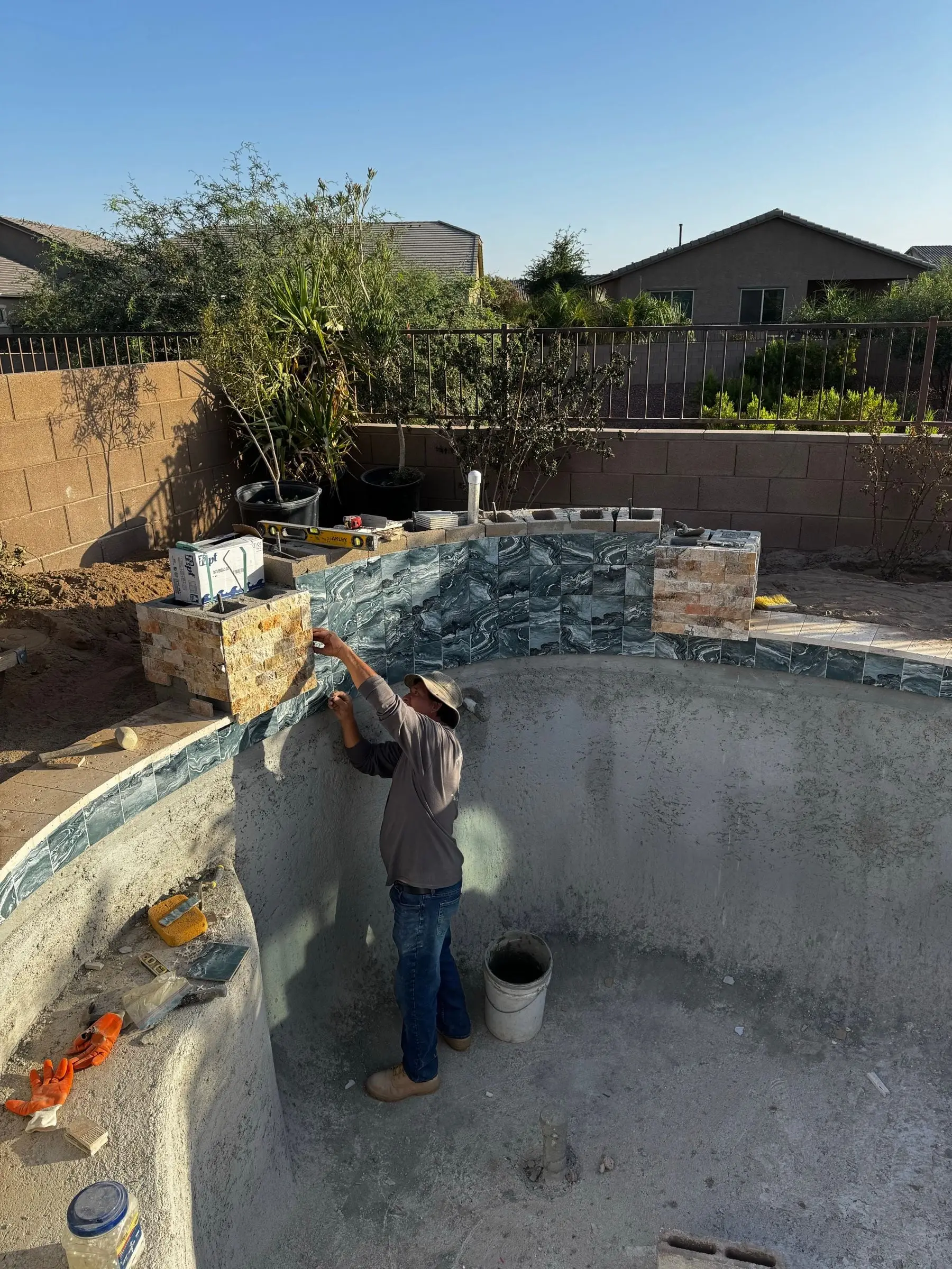 Tile setter installing stone-look mosaic tile on the raised wall of a freeform pool, Marana, AZ.