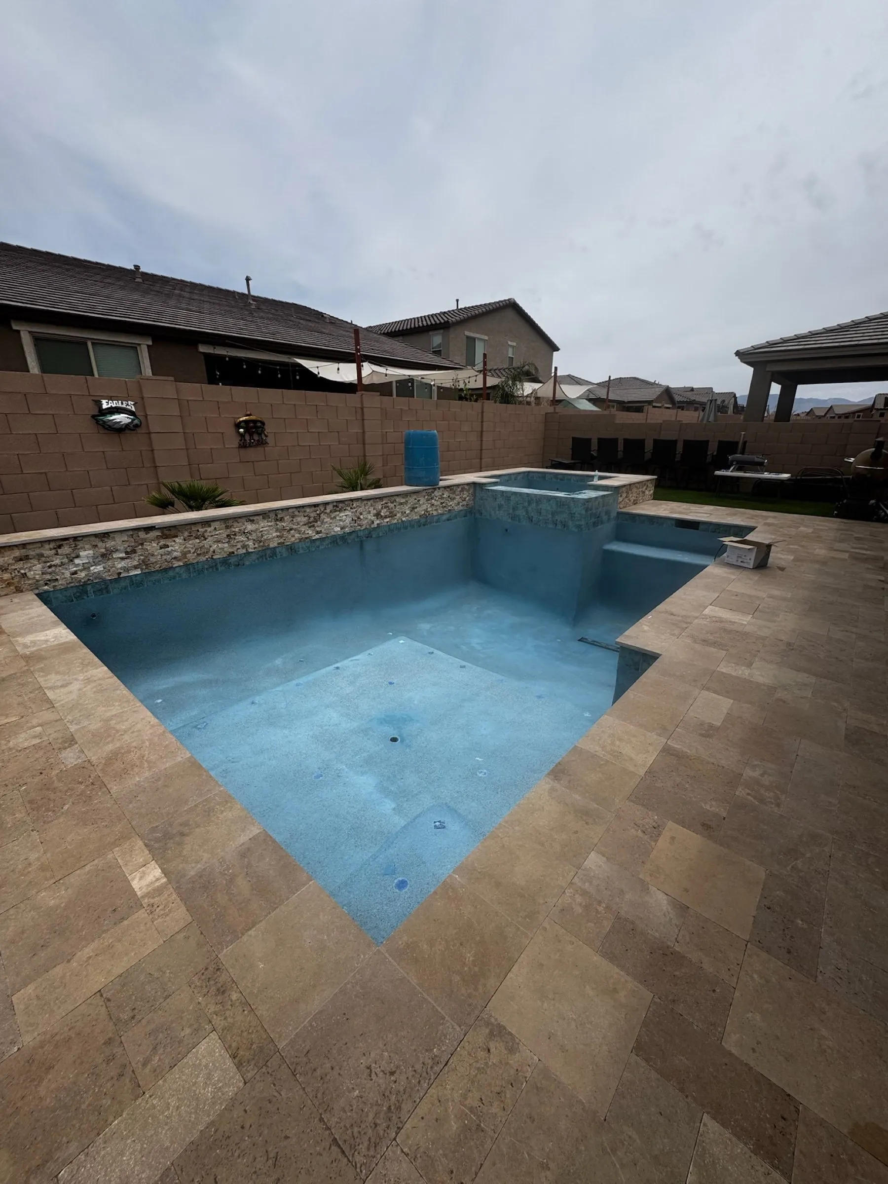 Newly filled pool with raised spa, stacked stone wall and walnut travertine deck under an overcast sky in Marana.