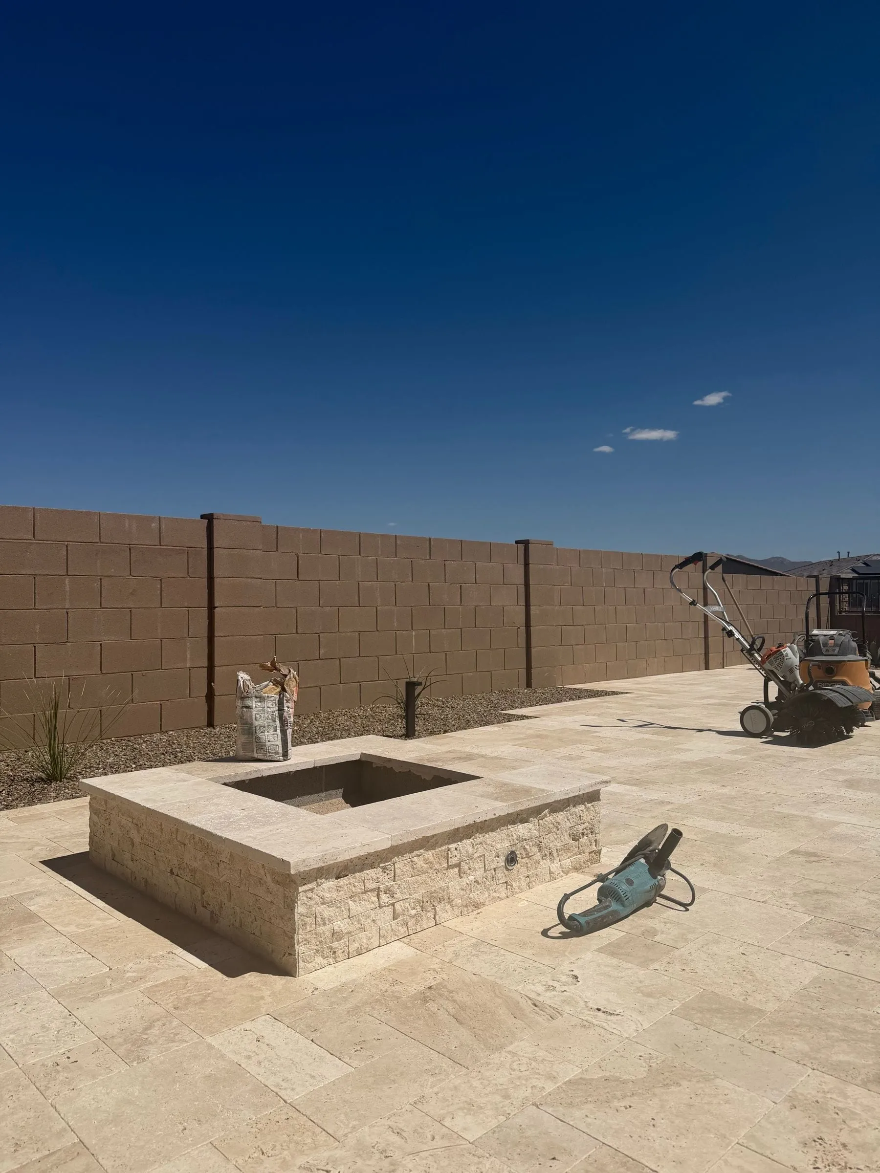 Square fire pit clad in stacked travertine on a new travertine paver patio, with desert plants along the wall.