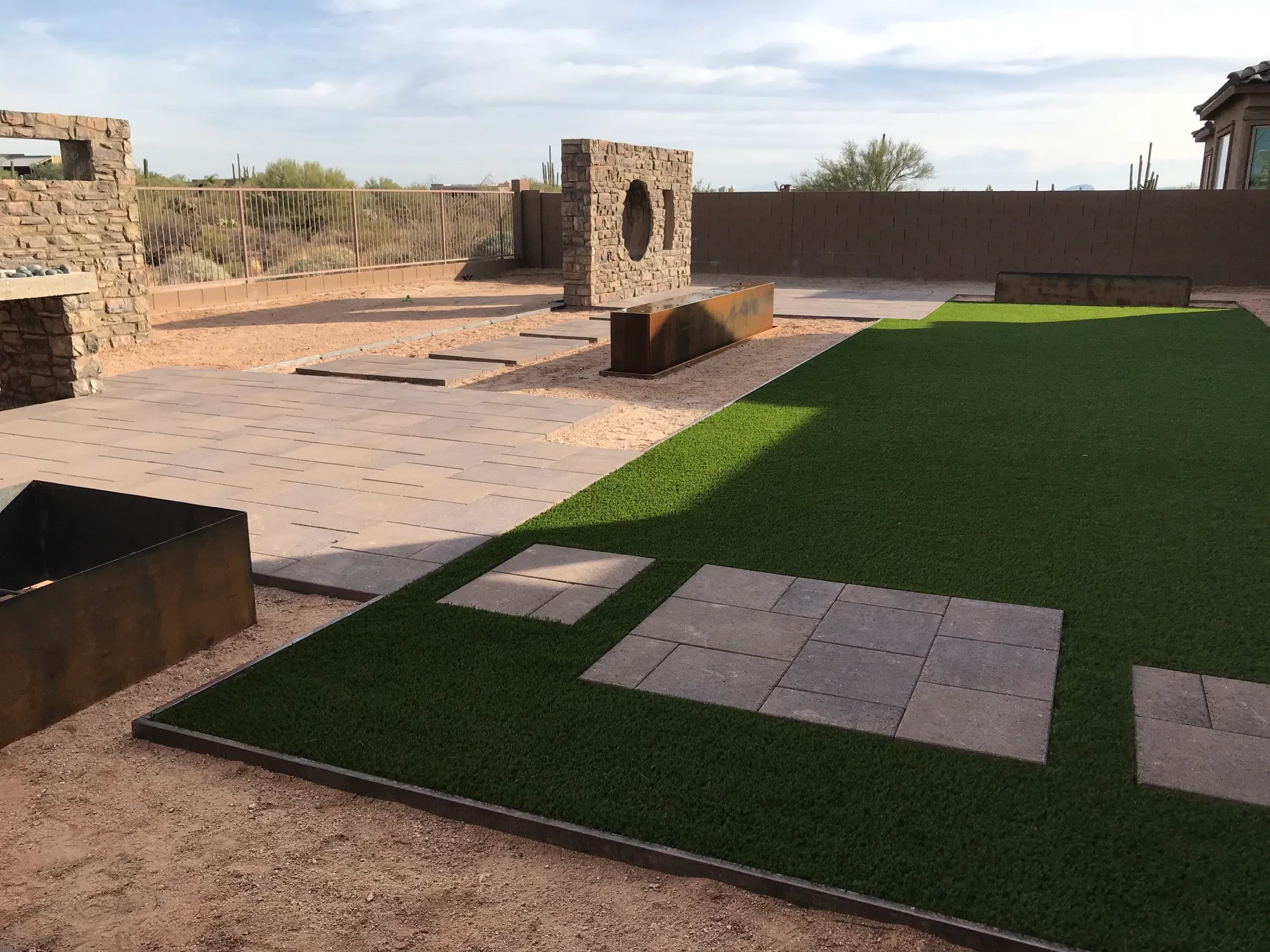 Backyard with artificial turf, paver pads, a stone wall feature and steel planters.